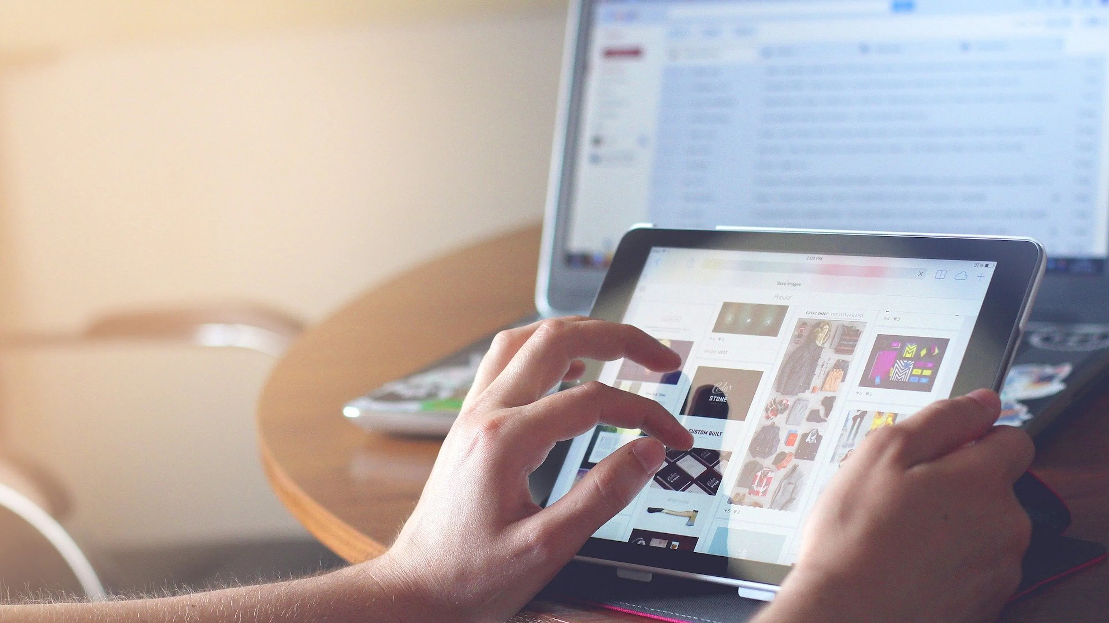 Close up of a persons hands tapping apps on an iPad with a laptop in the background