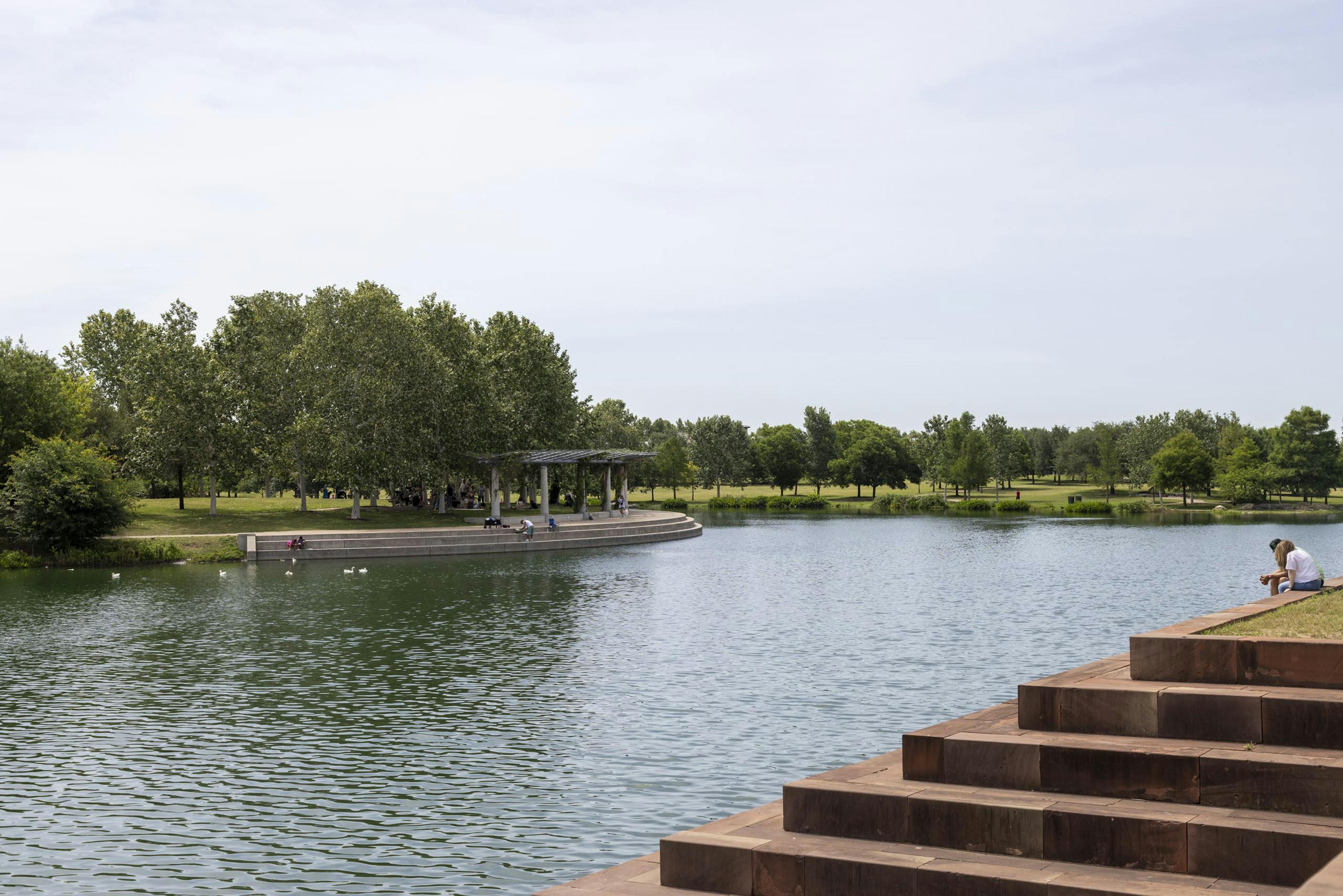 Mueller Lake during the day being enjoyed by park visitors feeding the ducks and sitting in the shade of a white pavilion