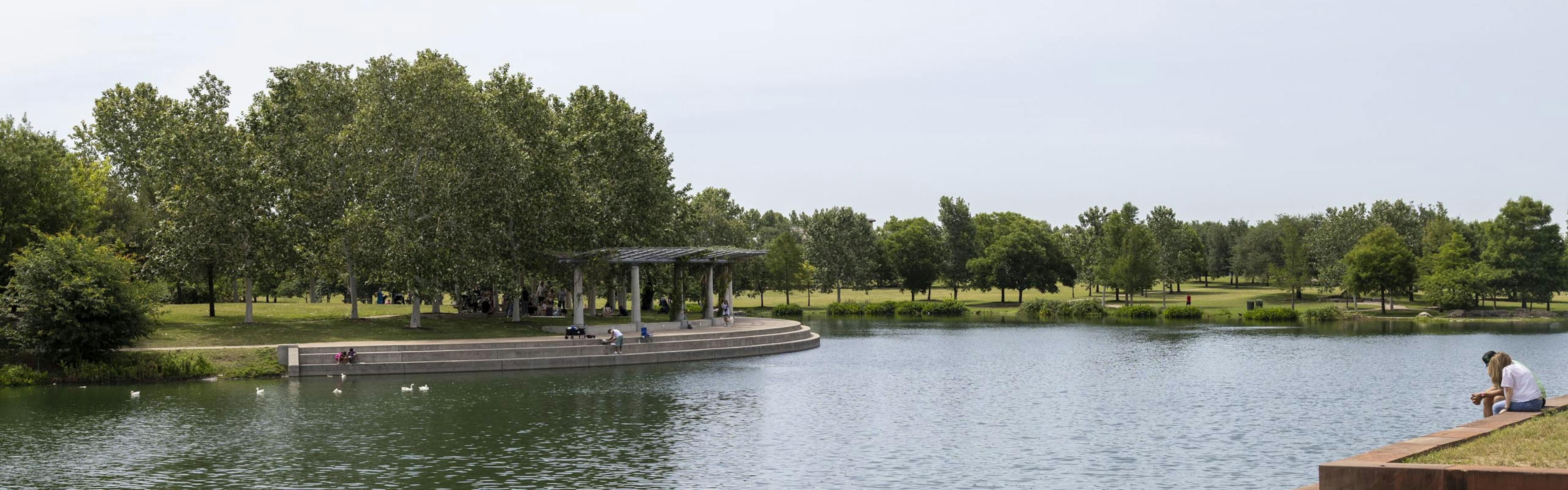 Mueller Lake during the day being enjoyed by park visitors feeding the ducks and sitting in the shade of a white pavilion