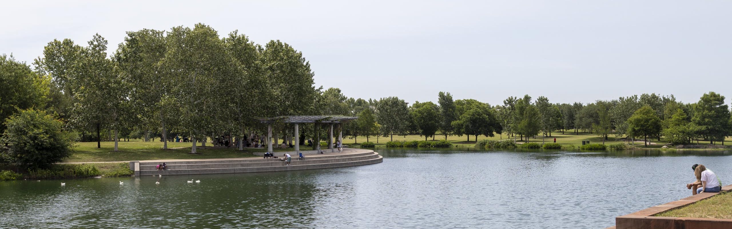 Mueller Lake during the day being enjoyed by park visitors feeding the ducks and sitting in the shade of a white pavilion