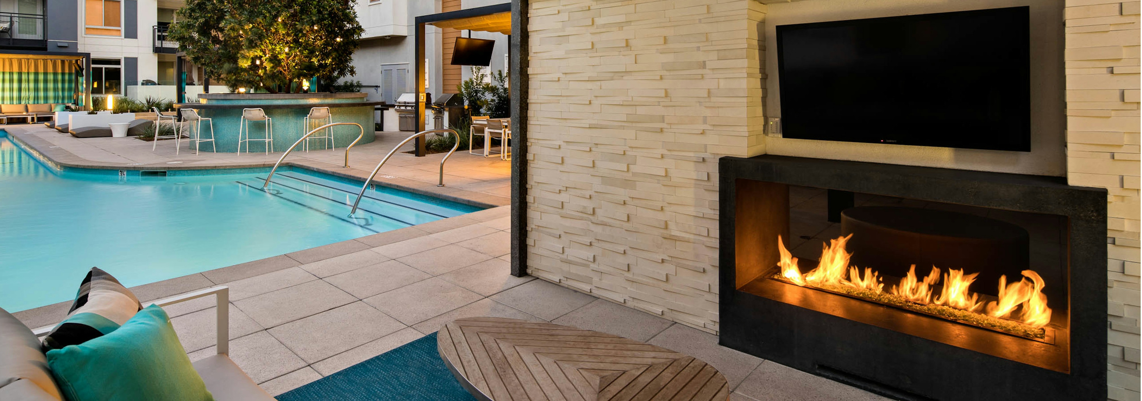 Dusk view of AMLI Uptown Orange outdoor fireplace with individual large screen TV and comfortable seating overlooking pool