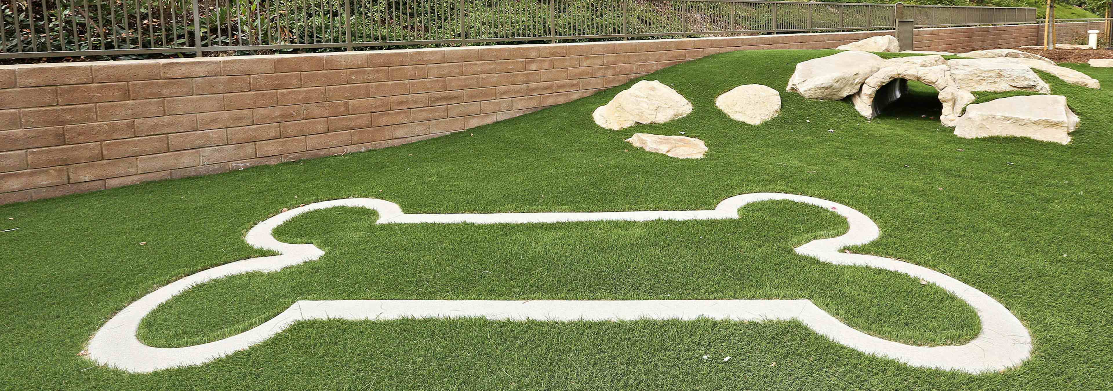 Exterior daytime close-up of pet park at AMLI Spanish Hills apartment with bark log tunnel and bone-shaped design on turf