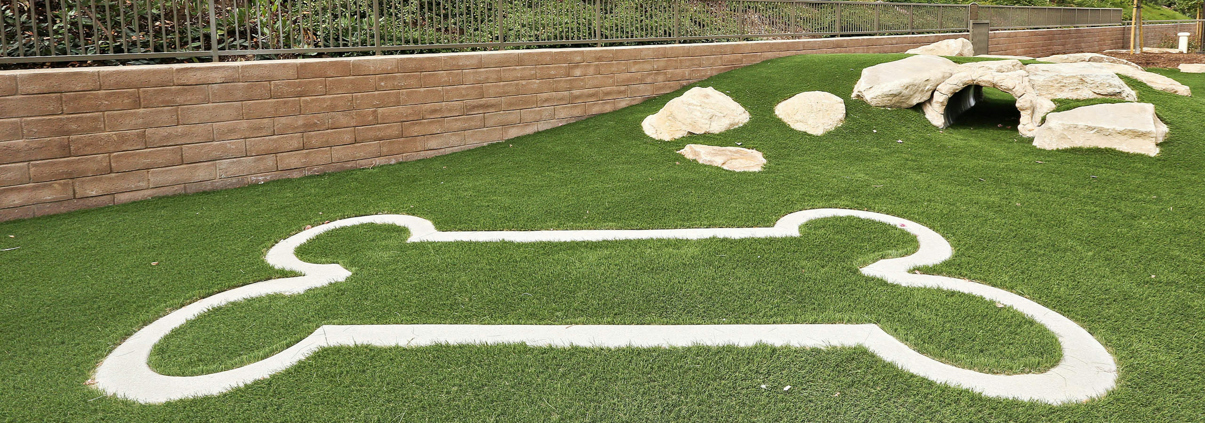 Exterior daytime close-up of pet park at AMLI Spanish Hills apartment with bark log tunnel and bone-shaped design on turf