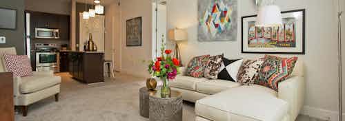 Living room at AMLI 5350 with carpet floors and colorful throw pillows on a white sofa with peek into kitchen in background