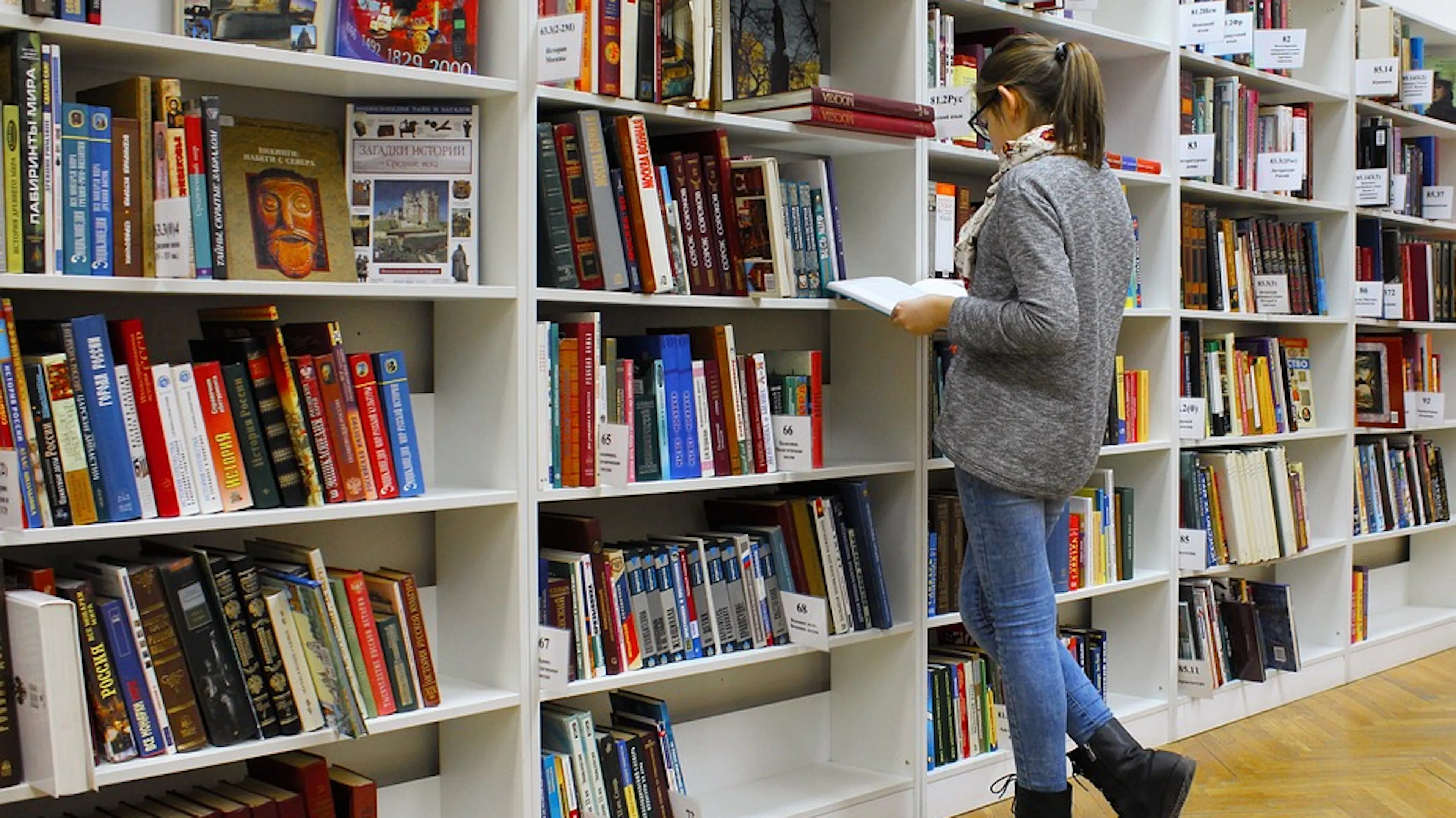 A woman facing a library bookshelf holding an open book in her hands and reading it