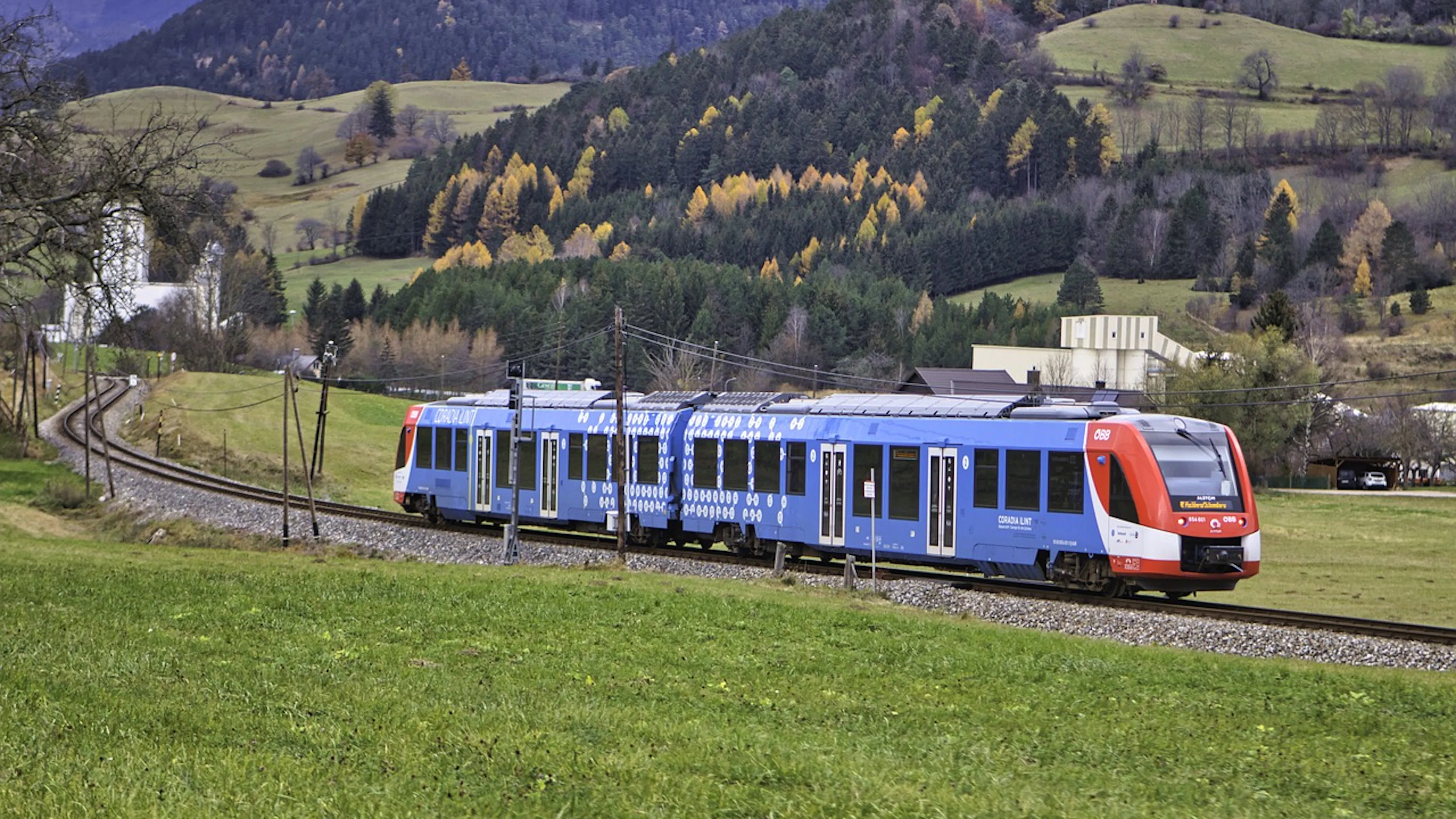 A two carriage train winding through a mountainous countryside with green meadows and forests