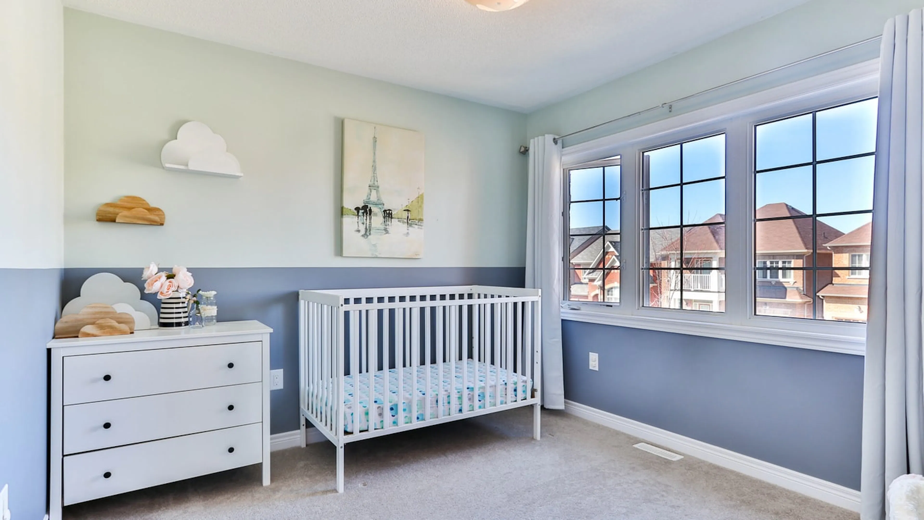 A blue and white nursery with a crib a dresser and some pale curtains