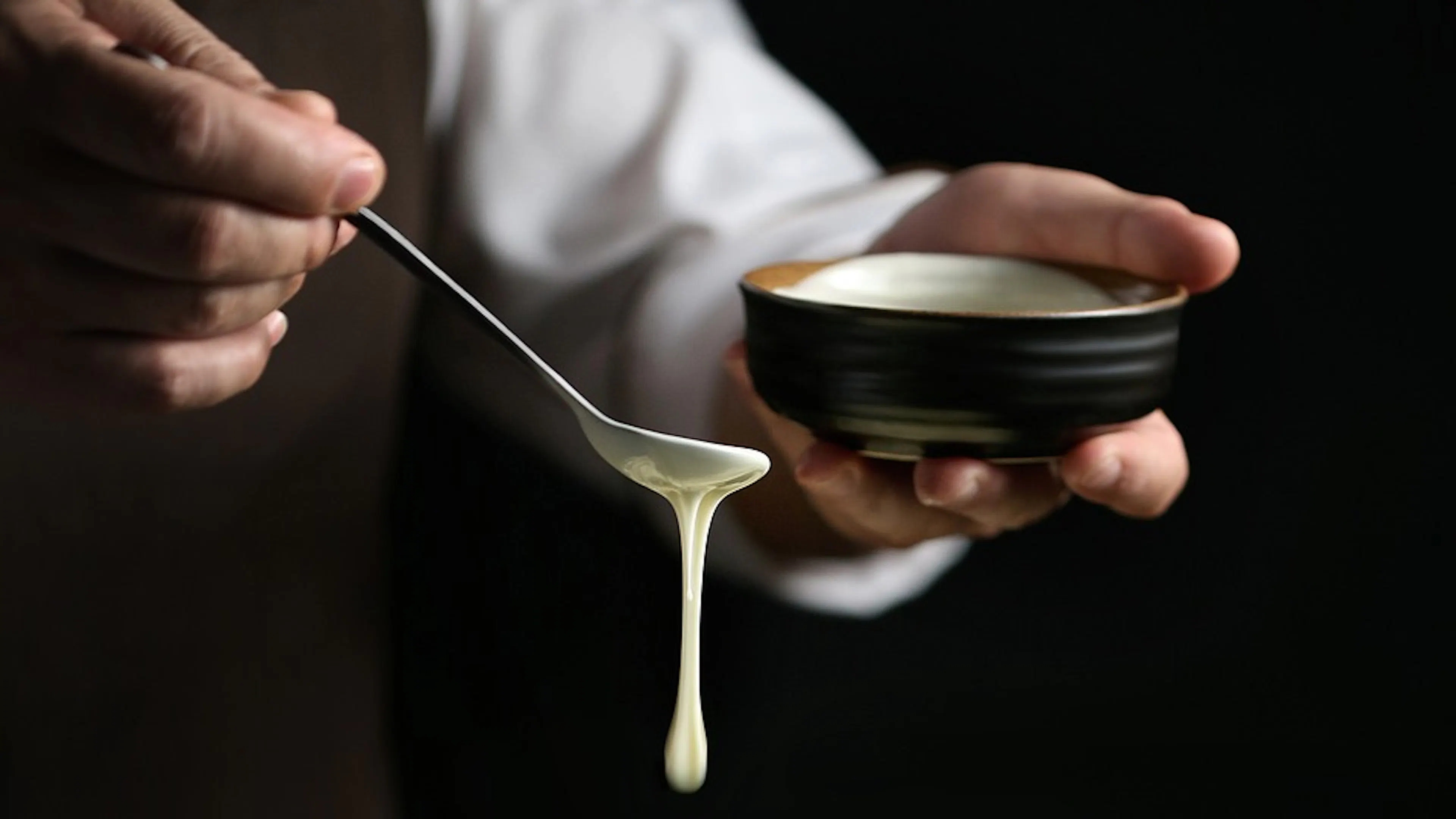 A chef spooning dripping melted white chocolate out of a small bowl using a silver spoon