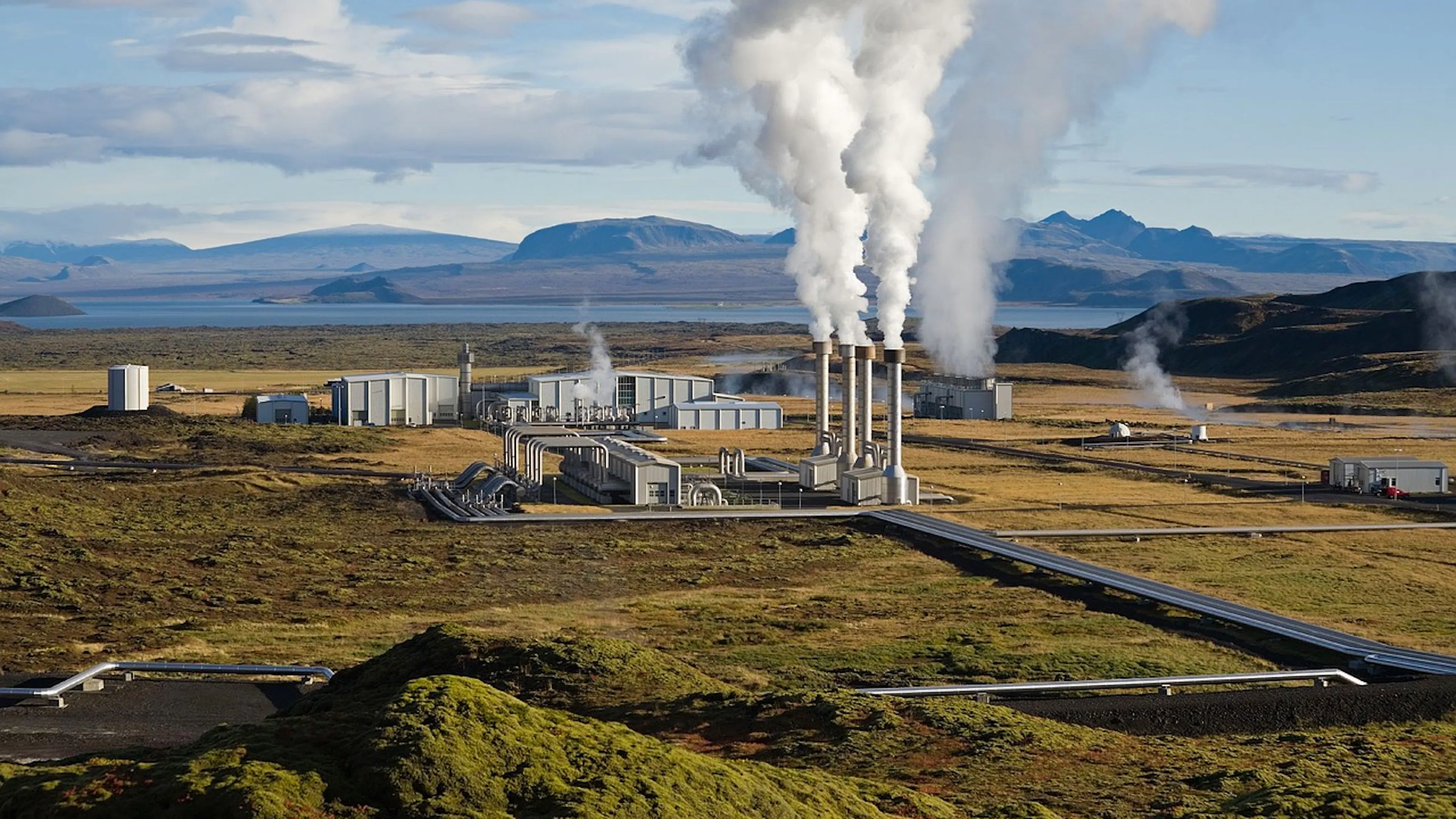 A scenic landscape of mountains and an inlet with a geothermal energy plant pumling steam into the air in the center of a valley