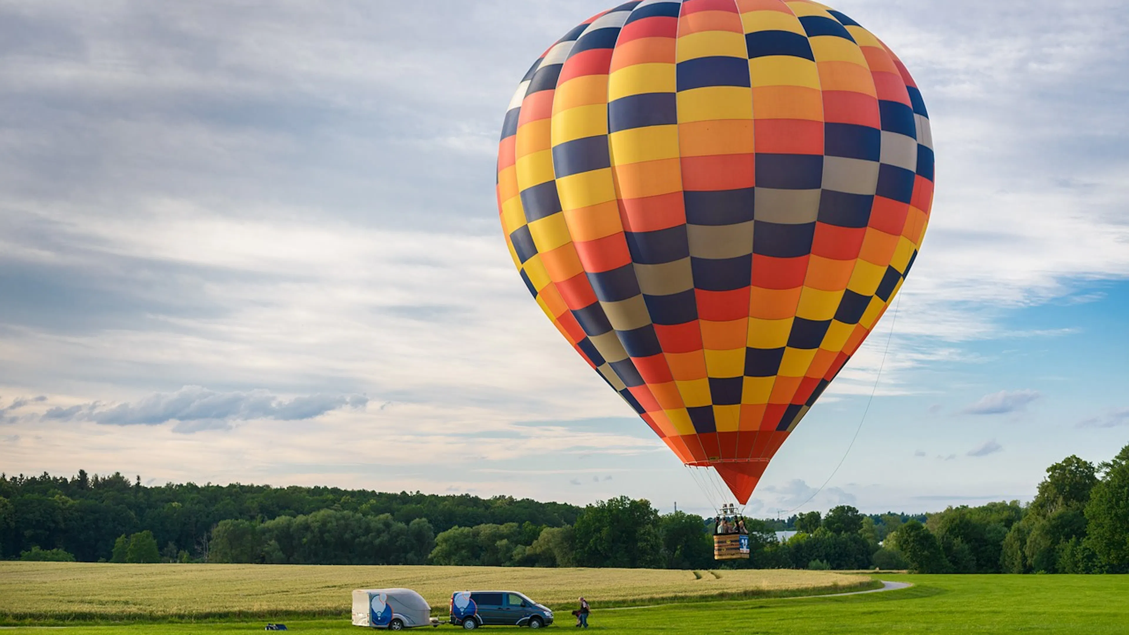 multi colored checked hot air balloon in front of blue sky over green field with van and trailer