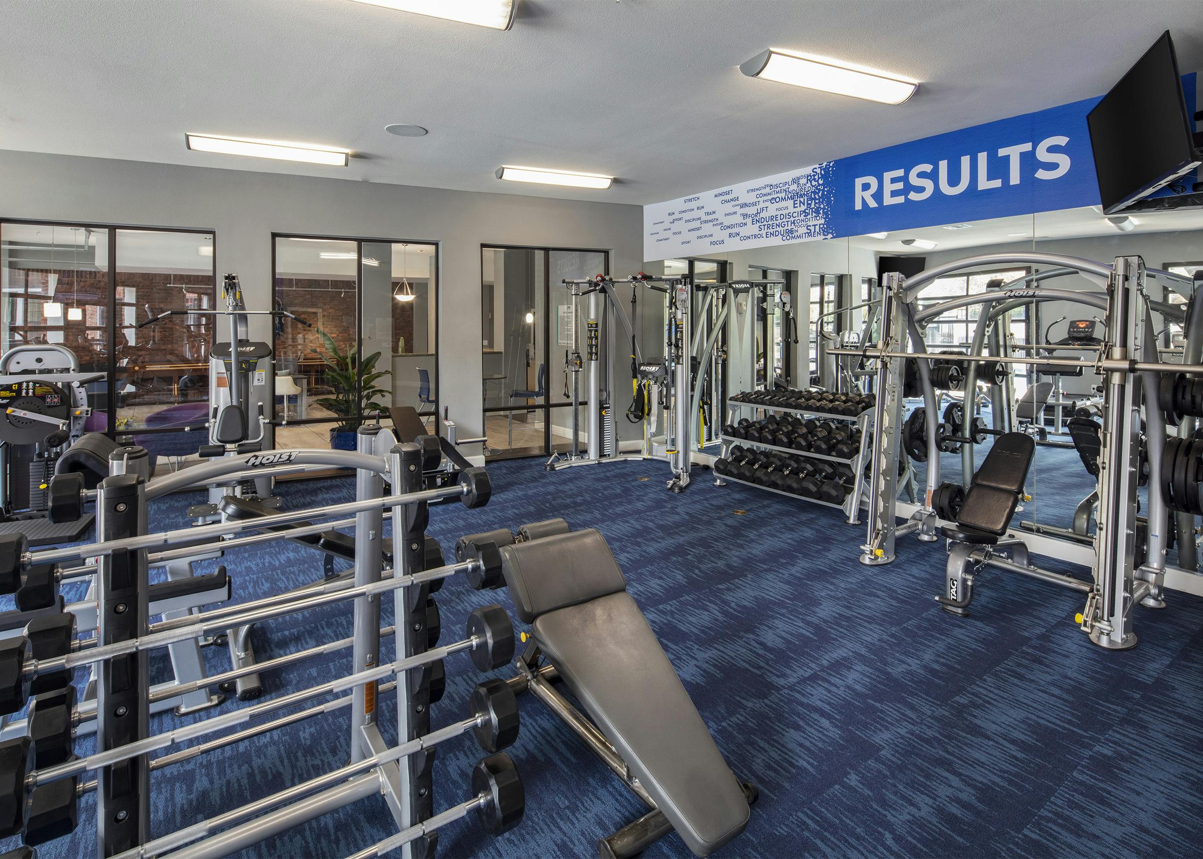 Fitness center at AMLI Eastside apartments with strength training machines and a dumbbell rack set on patterned blue carpet