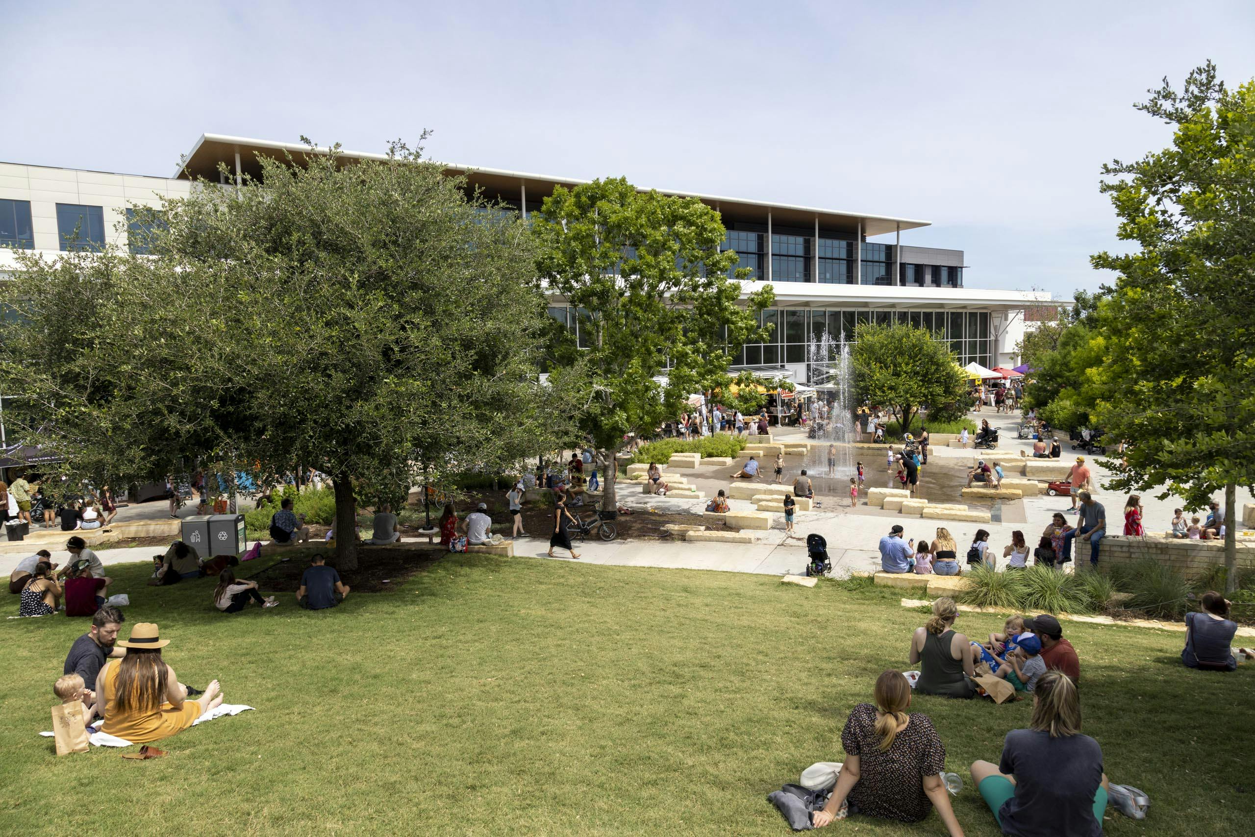 Branch Park Pavilion during the day featuring a lawn where families are picnicking and a water fountain feature with kids playing