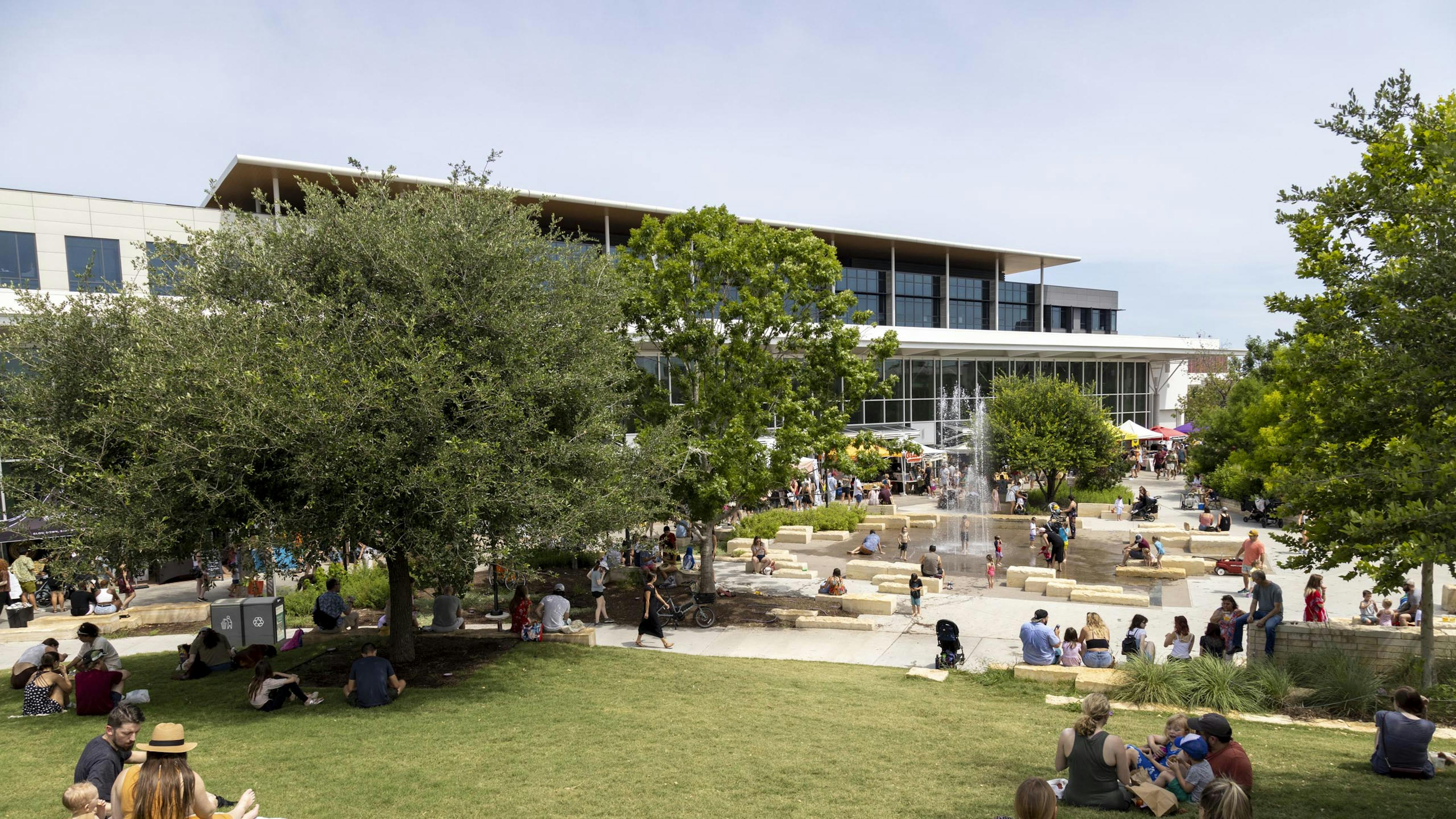 Branch Park Pavilion during the day featuring a lawn where families are picnicking and a water fountain feature with kids playing