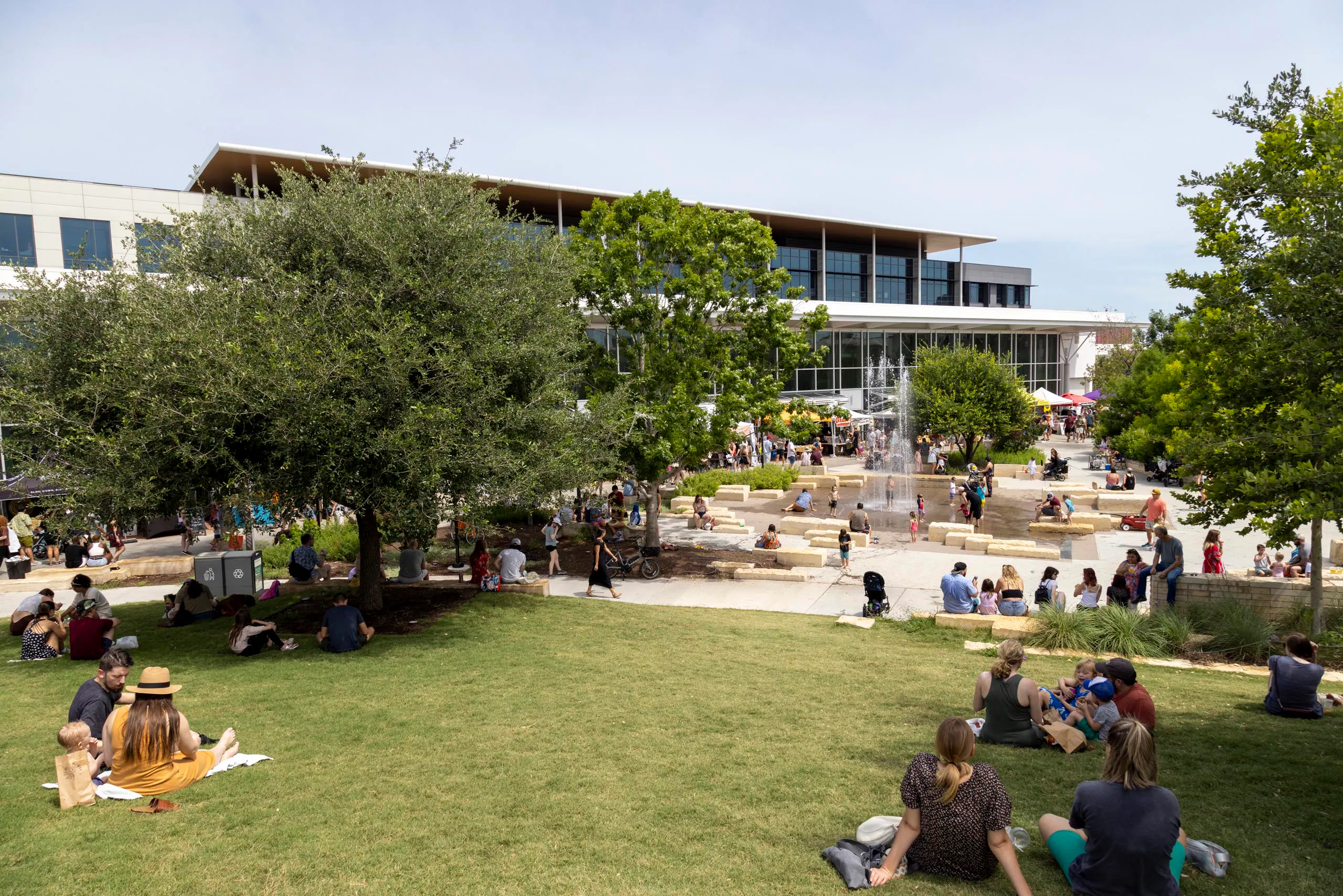 Branch Park Pavilion during the day featuring a lawn where families are picnicking and a water fountain feature with kids playing