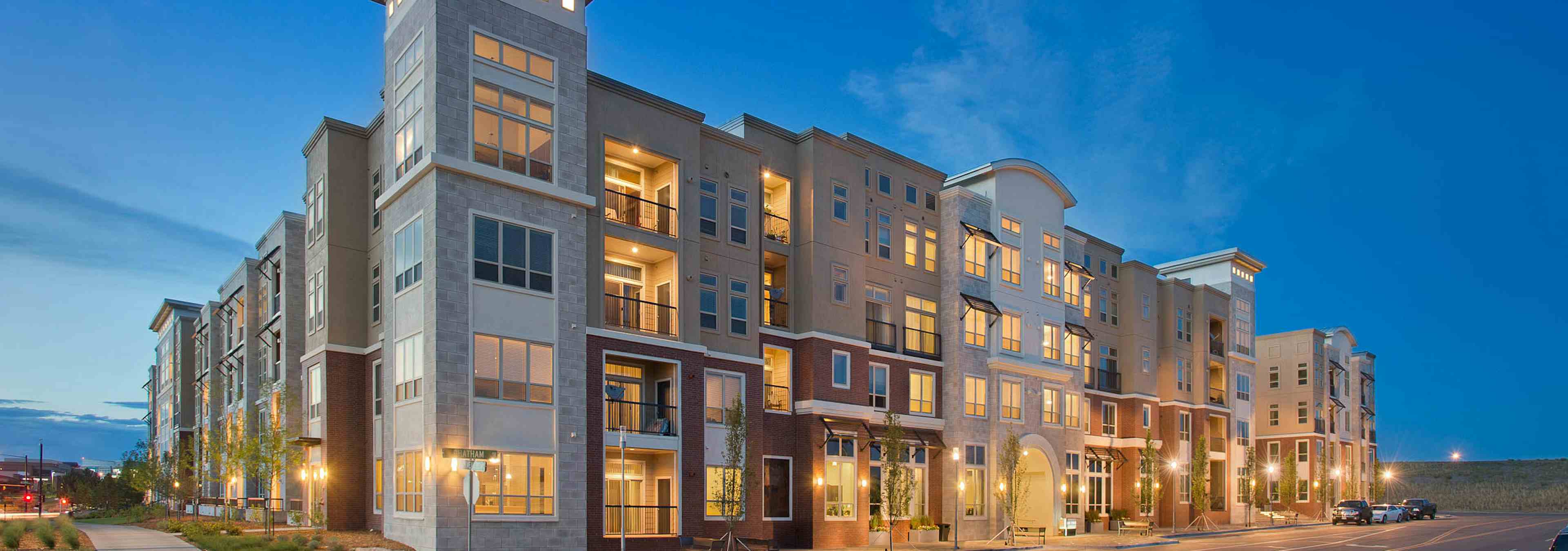 Exterior rendering of AMLI RidgeGate apartment building main entrance with lit windows and patios and trees and cars at dusk