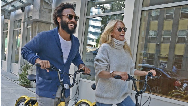 Close up of two people laughing as they walk their bikes along the sidewalk with large building windows in the background