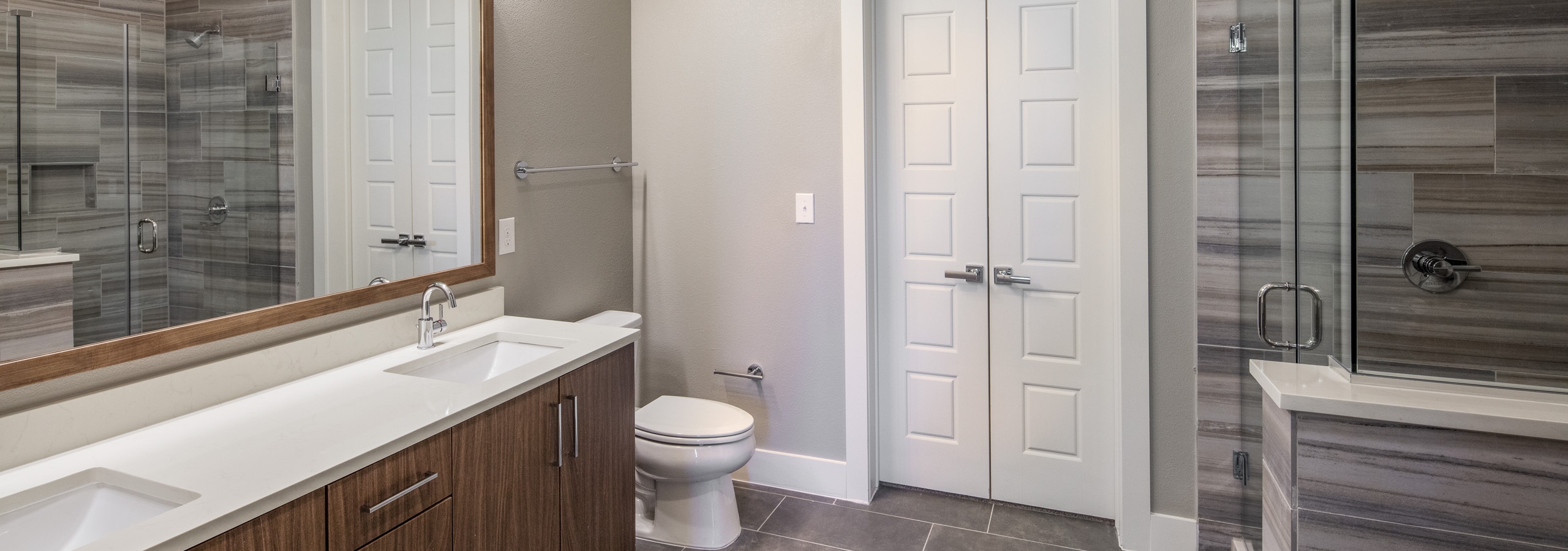 AMLI South Shore apartment bathroom with glass shower with tan tile surround and double vanity with brown wood cabinets