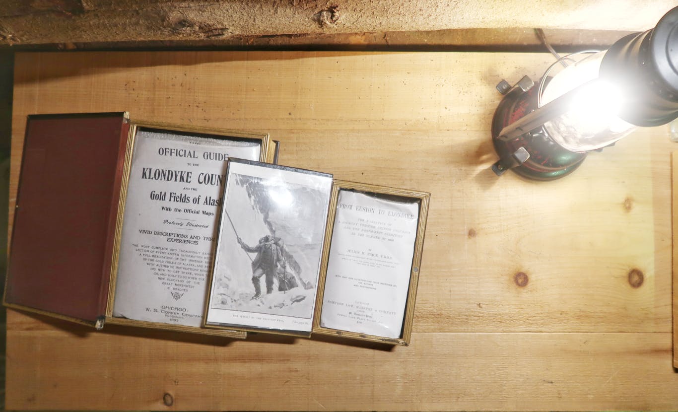 A display of old books with pictures about the Klondike Gold Rush set on a wooden table next to an oil lamp