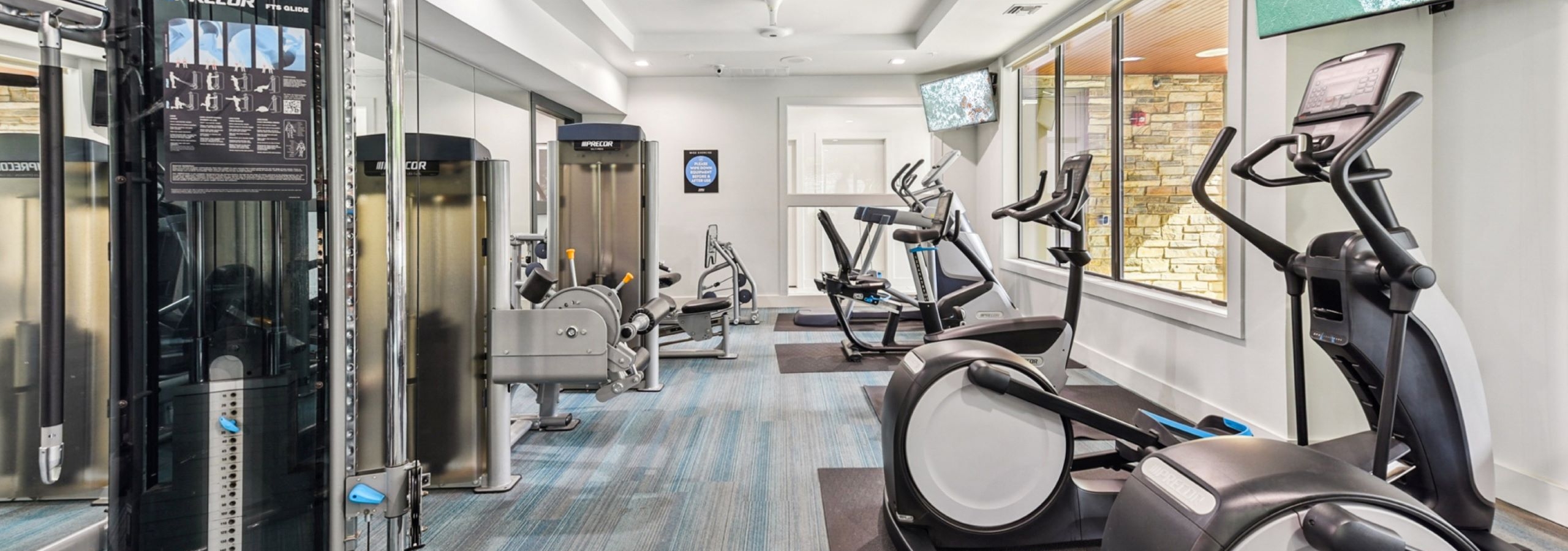 Modern fitness center at AMLI Covered Bridge apartments with various exercise equipment, large windows and flat-screen TVs