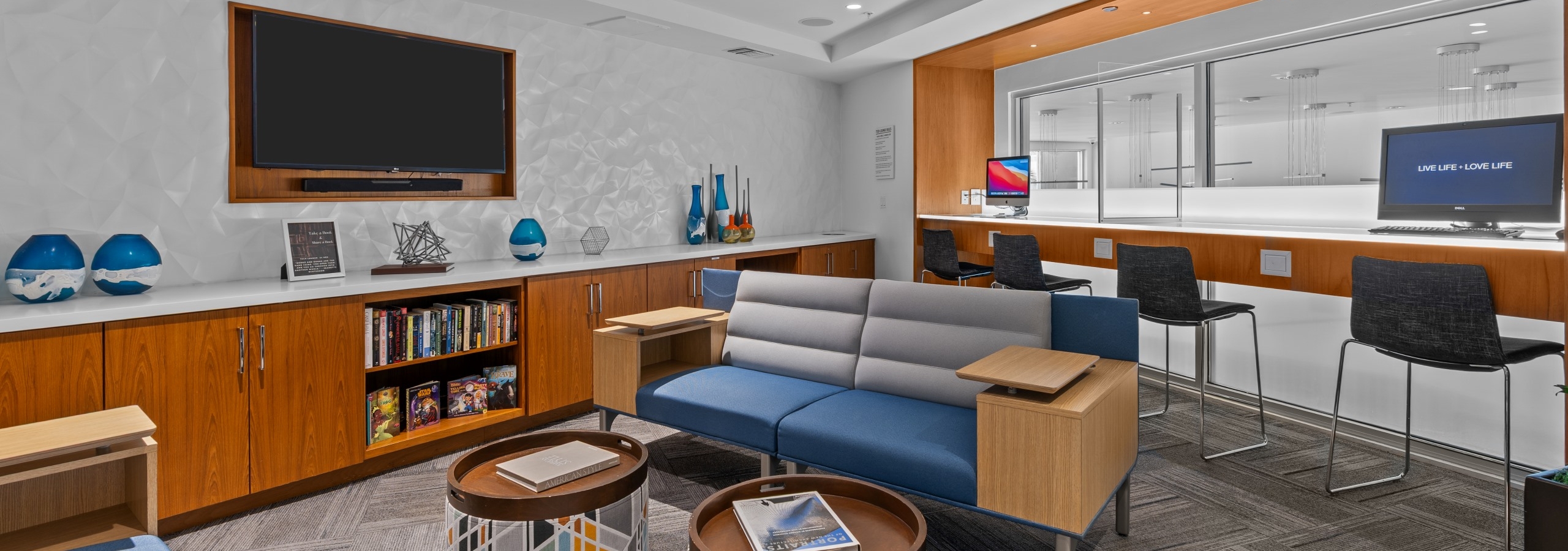 Tech lounge with two computers and various blue and gray seating and big screen TV at AMLI Uptown Orange apartments