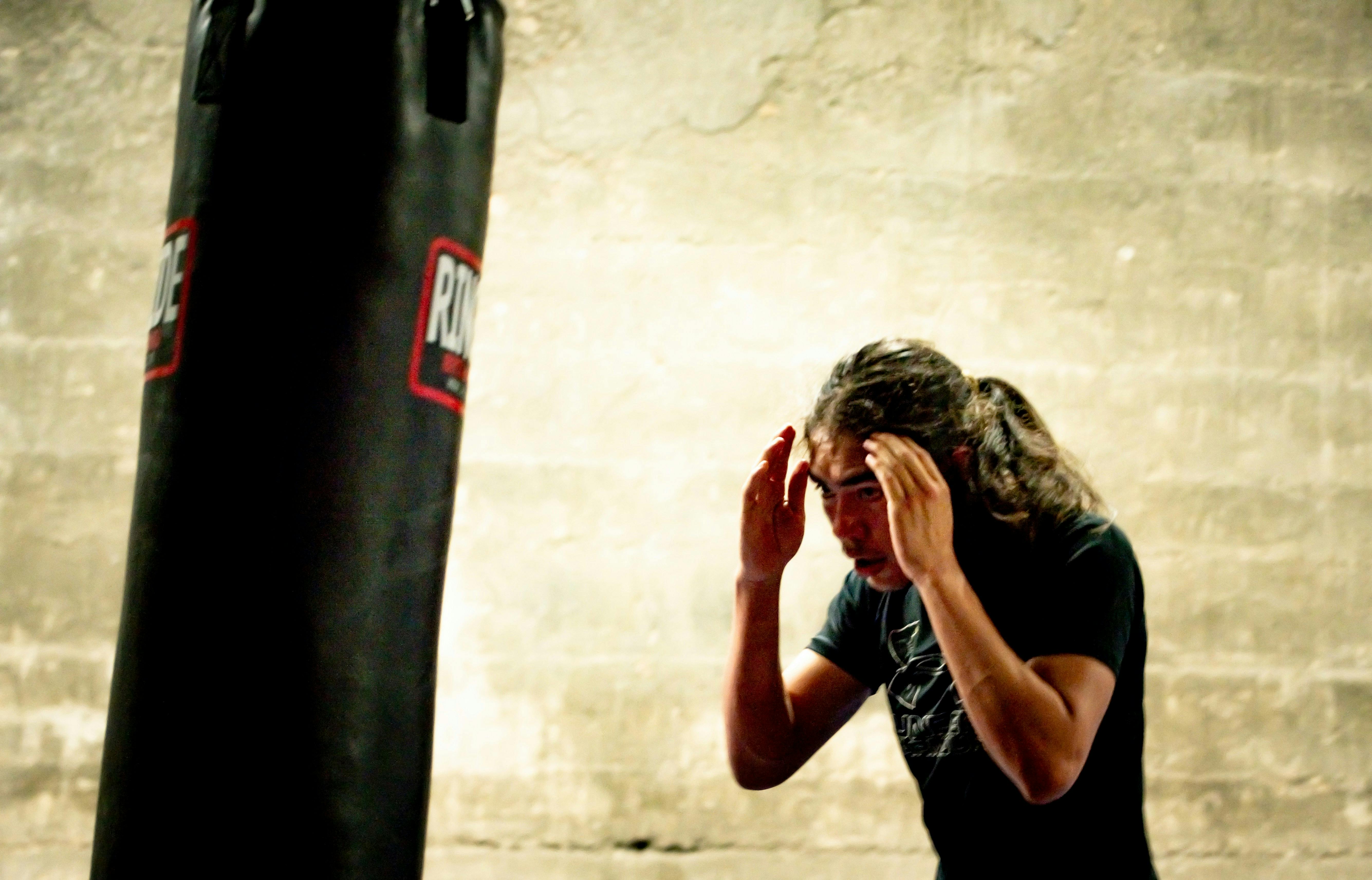 A person with long, dark hair pulled back, wearing a black t-shirt, stands facing a tall, black punching bag with red lettering. Their hands are raised near their head, seemingly in a defensive or focused stance.