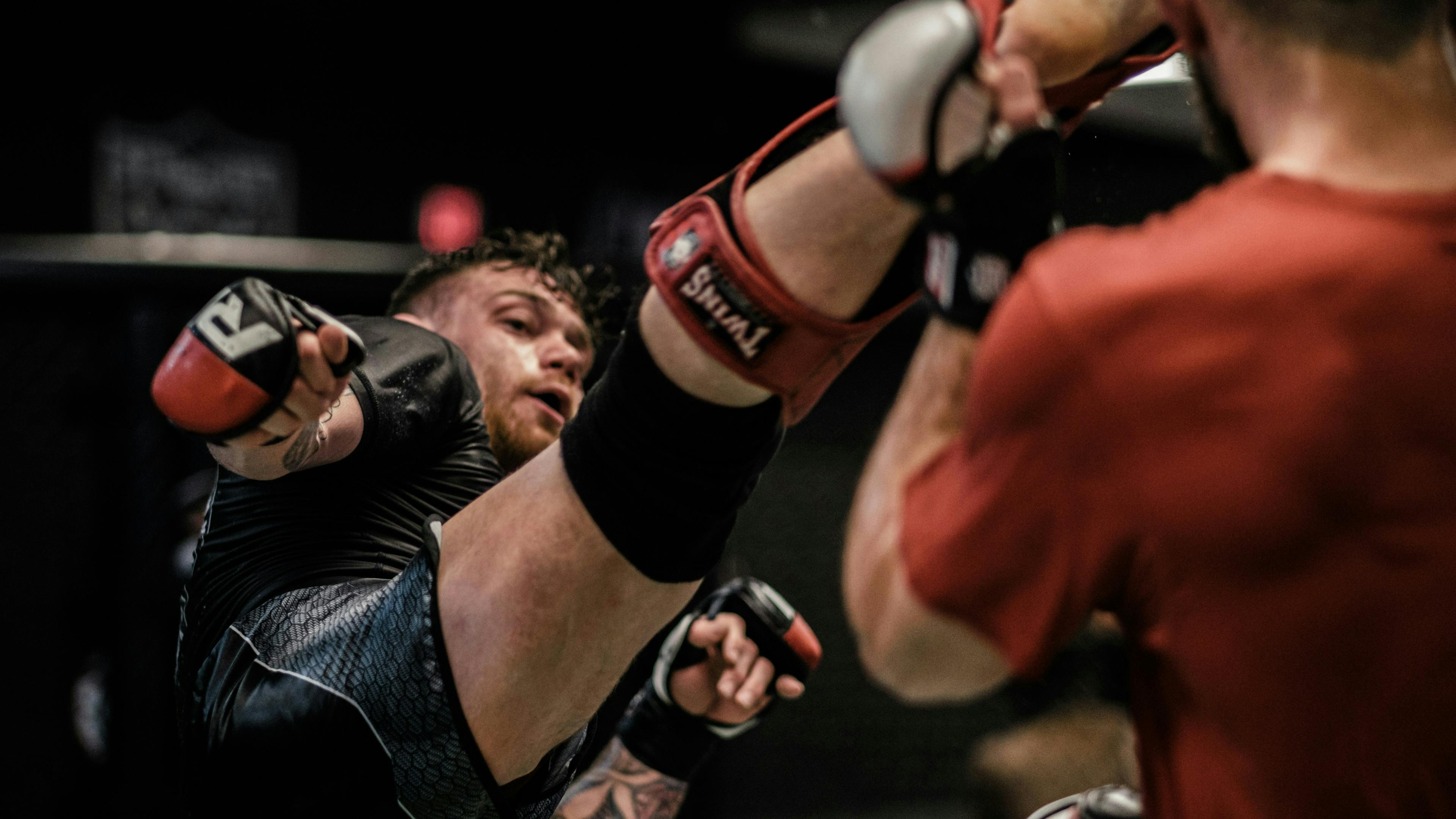 Two male mixed martial arts (MMA) fighters are sparring. The fighter in the foreground, wearing black and grey shorts and shin guards, is mid-kick with his leg extended towards his opponent, whose back is partially visible.