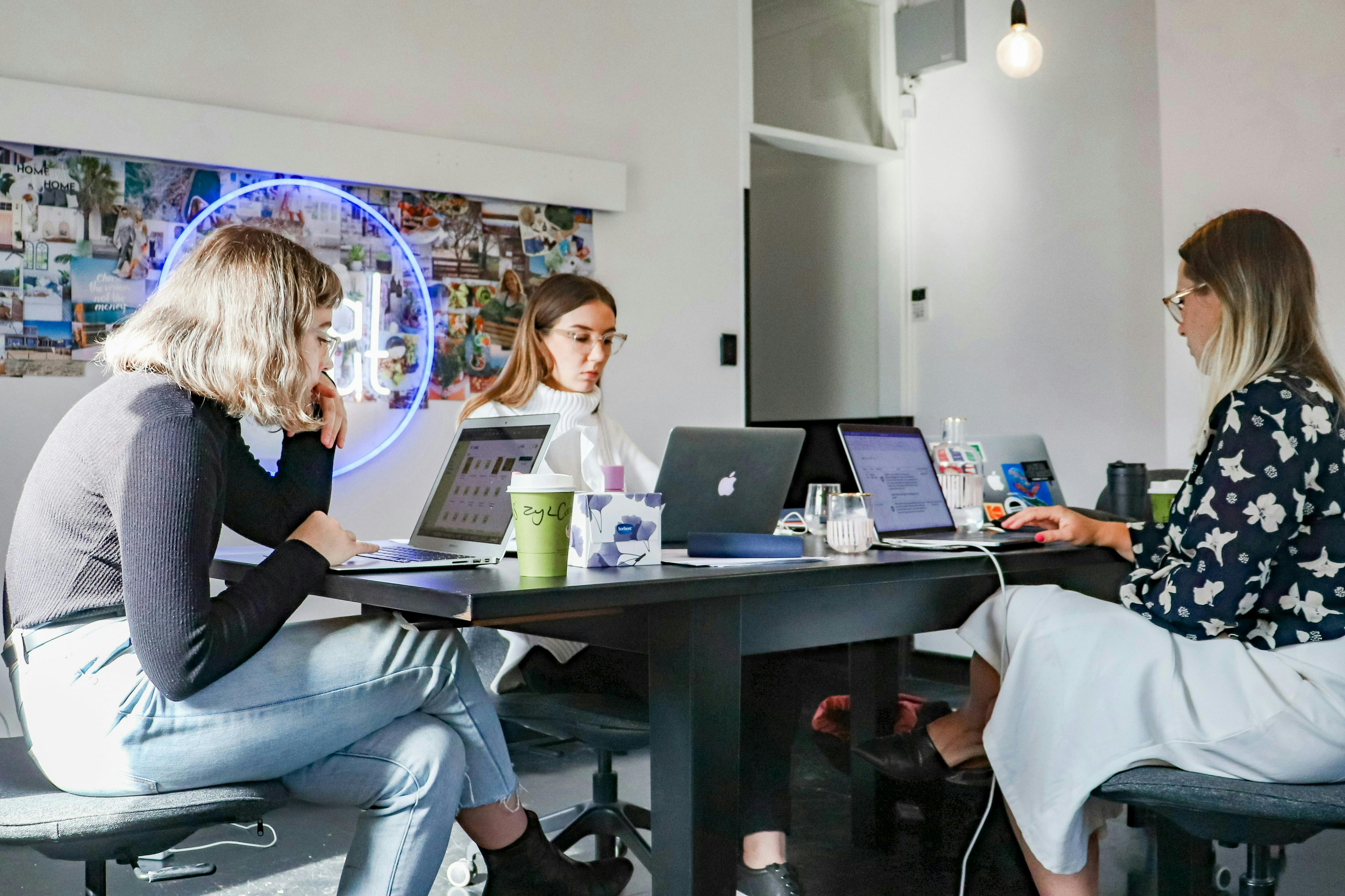 Three women work on laptops at a long black table in a modern office space. A bright blue neon sign glows on the wall behind them.
