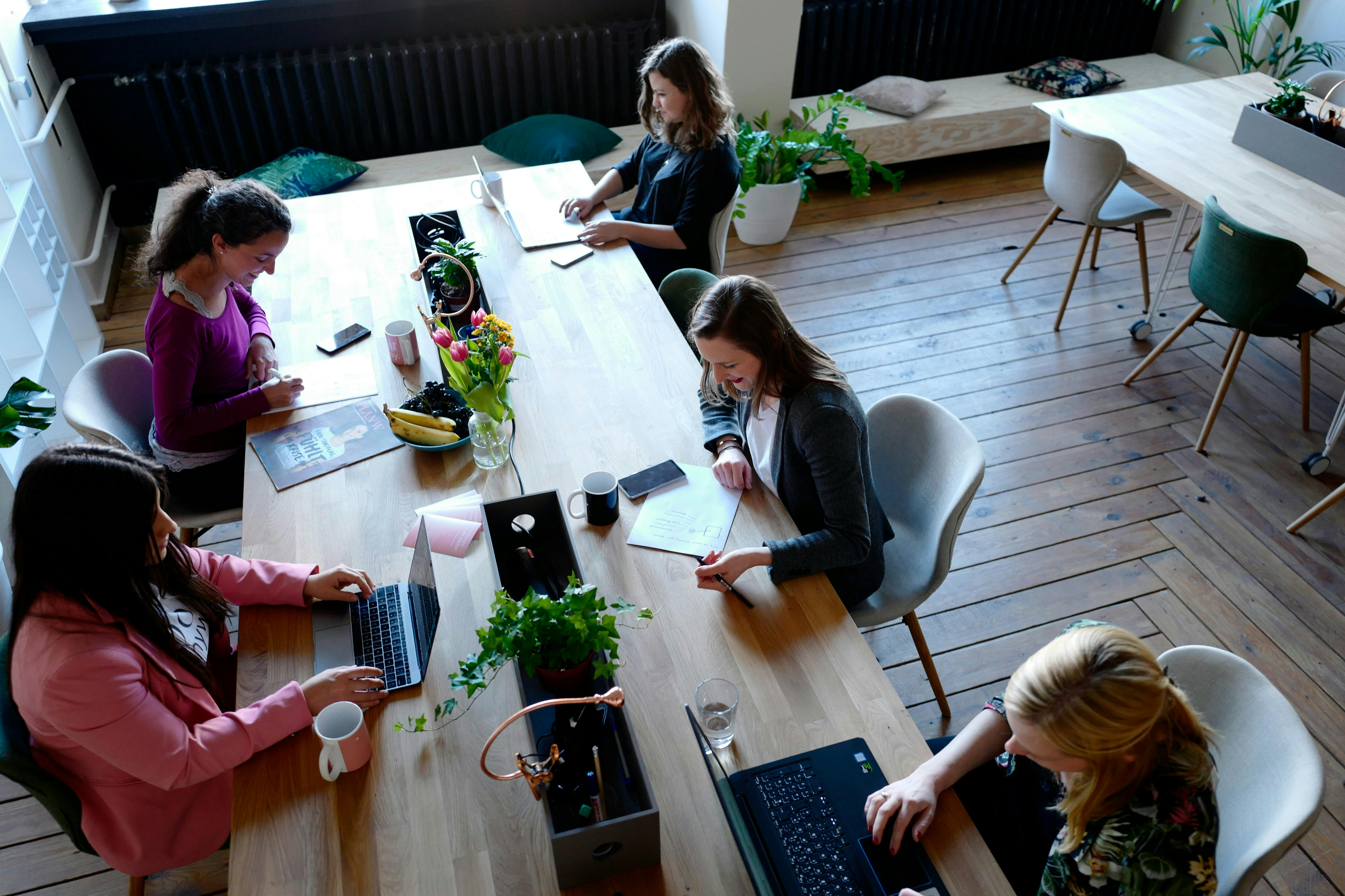 An overhead view of five women working on laptops and papers at a long wooden table in a brightly lit coworking space, with plants and natural light.