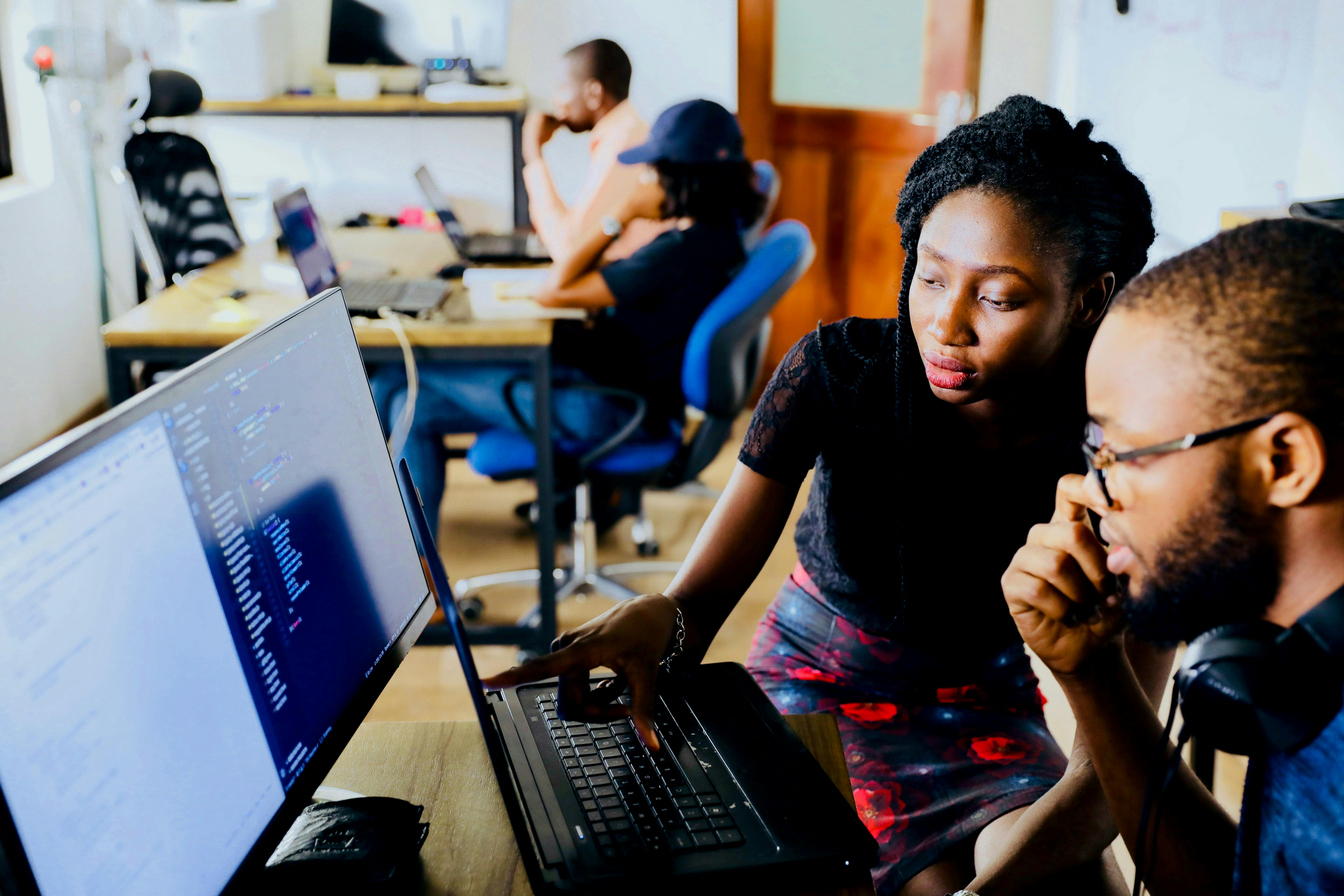 Two individuals, appearing to be of African descent, are focused on a laptop screen in an office environment. The person on the left points at the screen while the person on the right looks on, with other people working in the background.