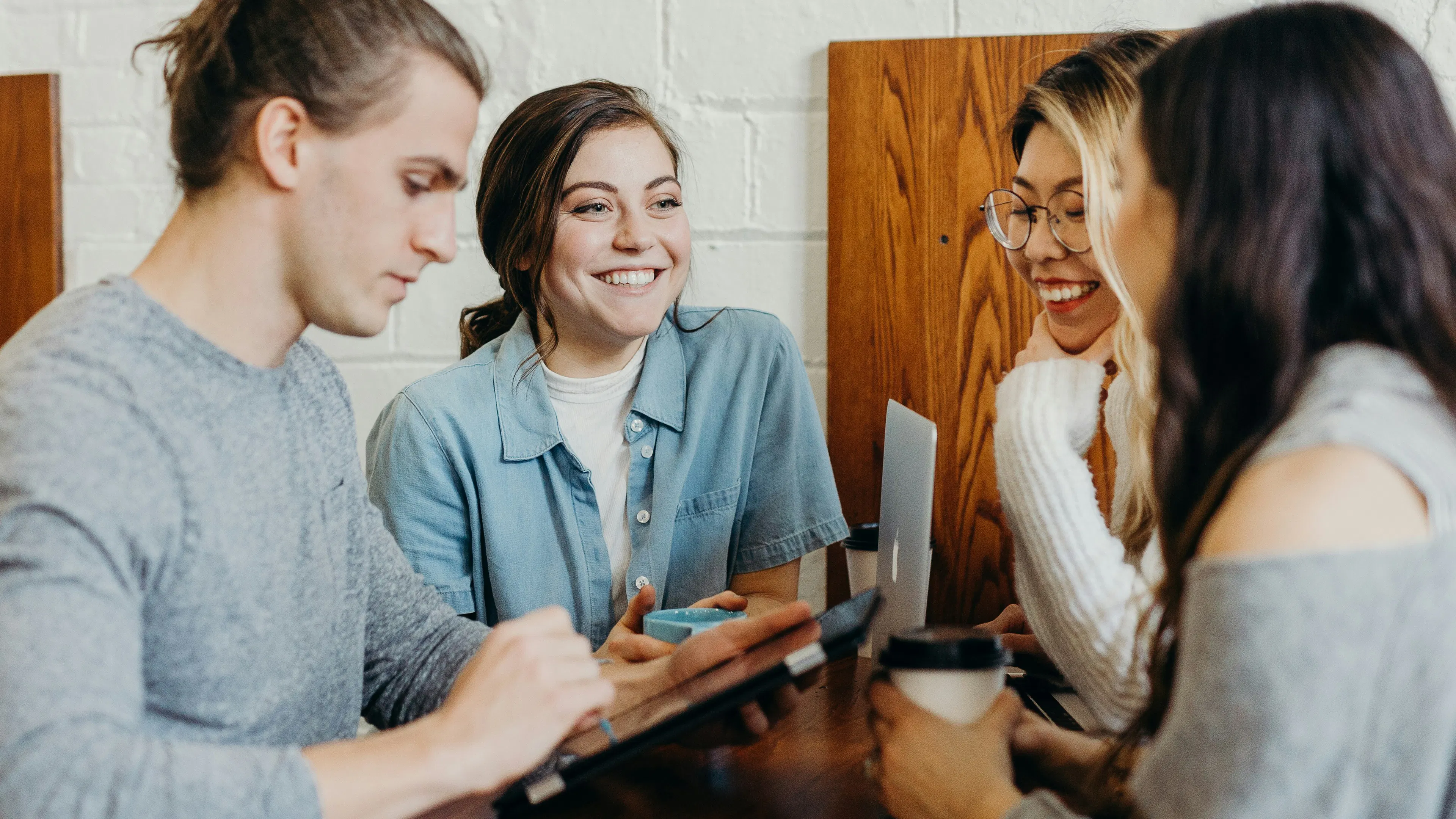 Four young adults are gathered around a wooden table. A man with a top knot on the left is looking at a tablet. A woman in a denim shirt smiles and looks towards the right. Two women on the right are also smiling, one with glasses holding a coffee cup.