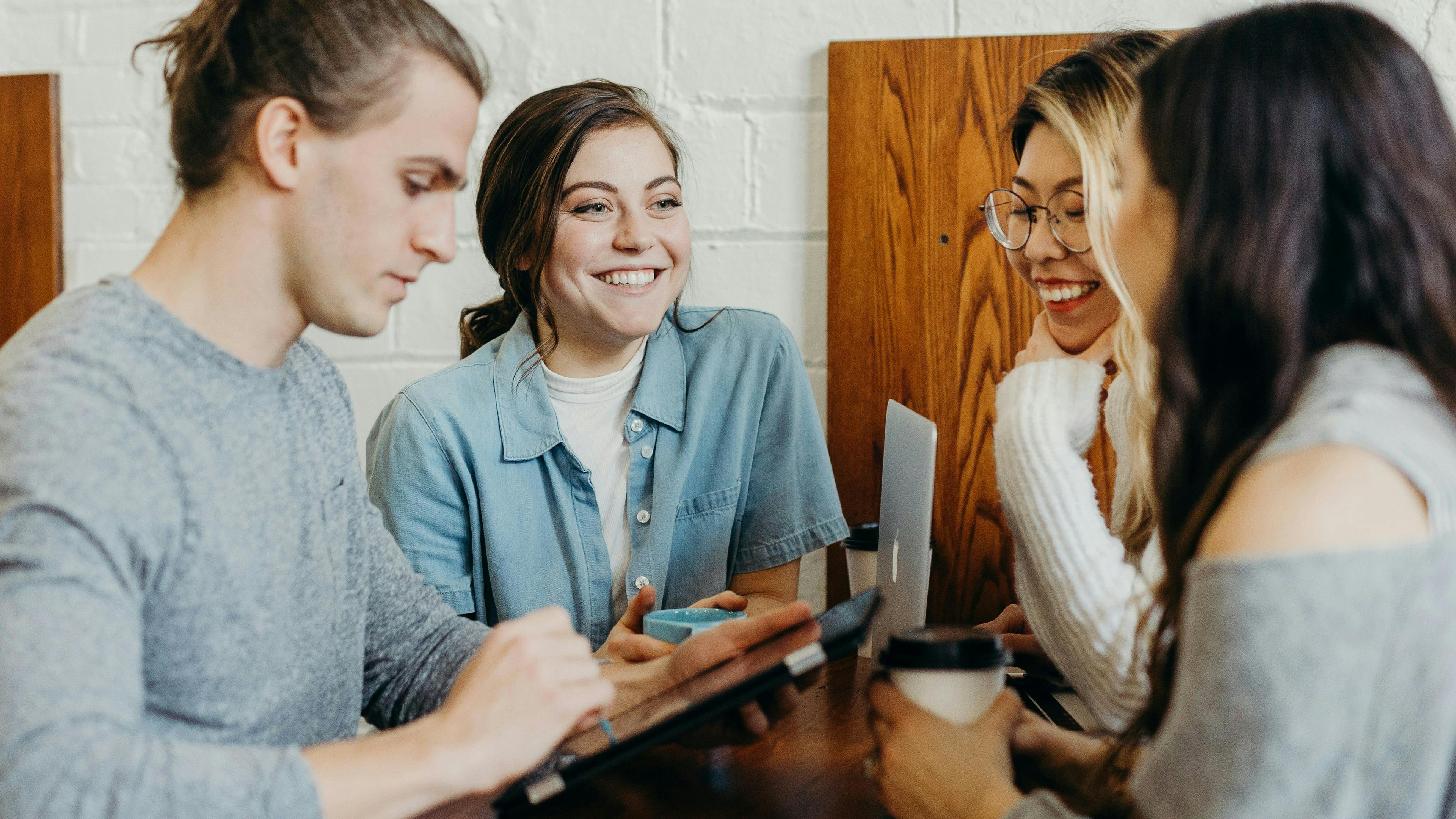 Four young adults are gathered around a wooden table. A man with a top knot on the left is looking at a tablet. A woman in a denim shirt smiles and looks towards the right. Two women on the right are also smiling, one with glasses holding a coffee cup.