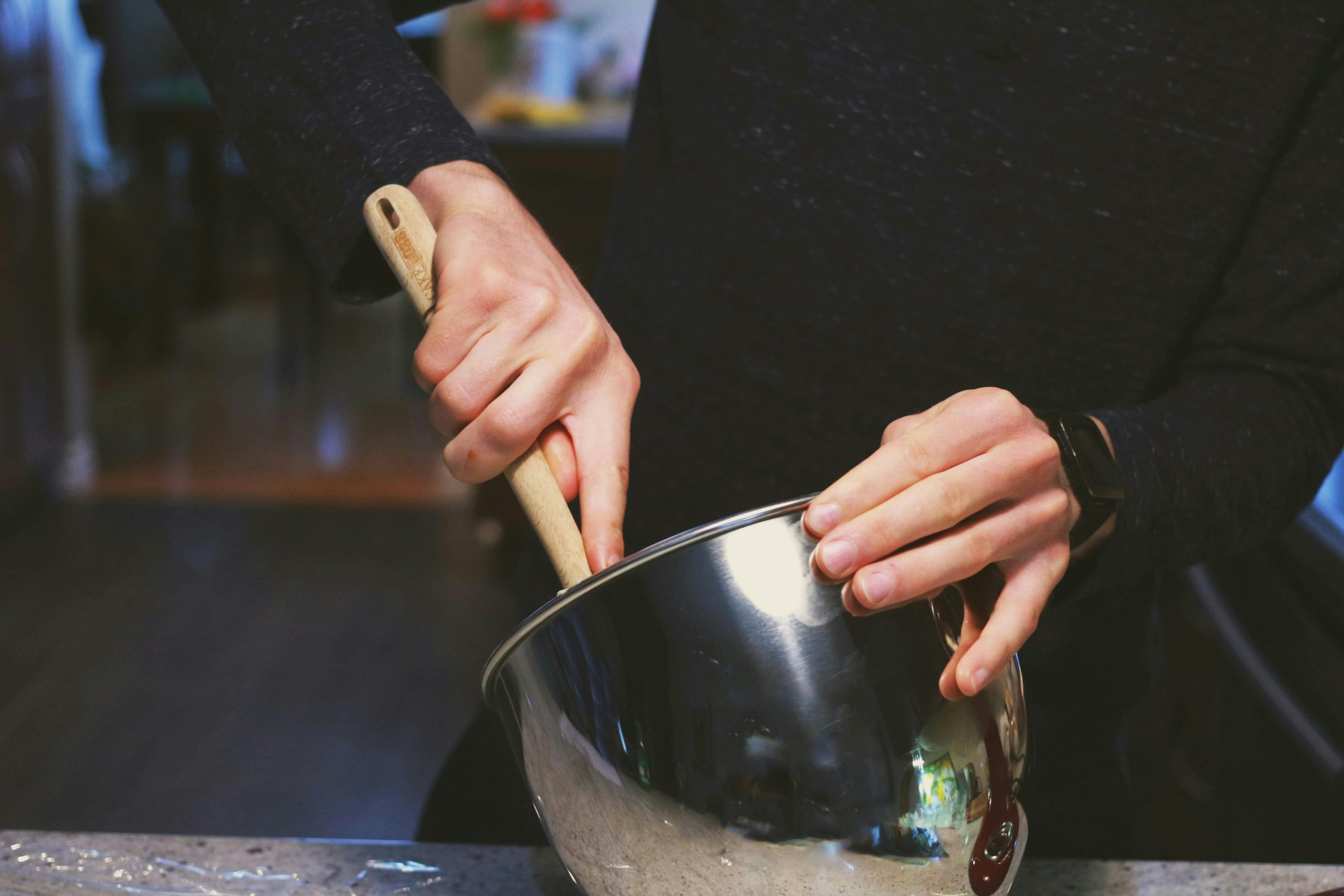 A person, wearing a long-sleeved dark shirt, uses a wooden spoon to mix ingredients in a large, shiny stainless steel mixing bowl. Their hands are holding the spoon and steadying the bowl.