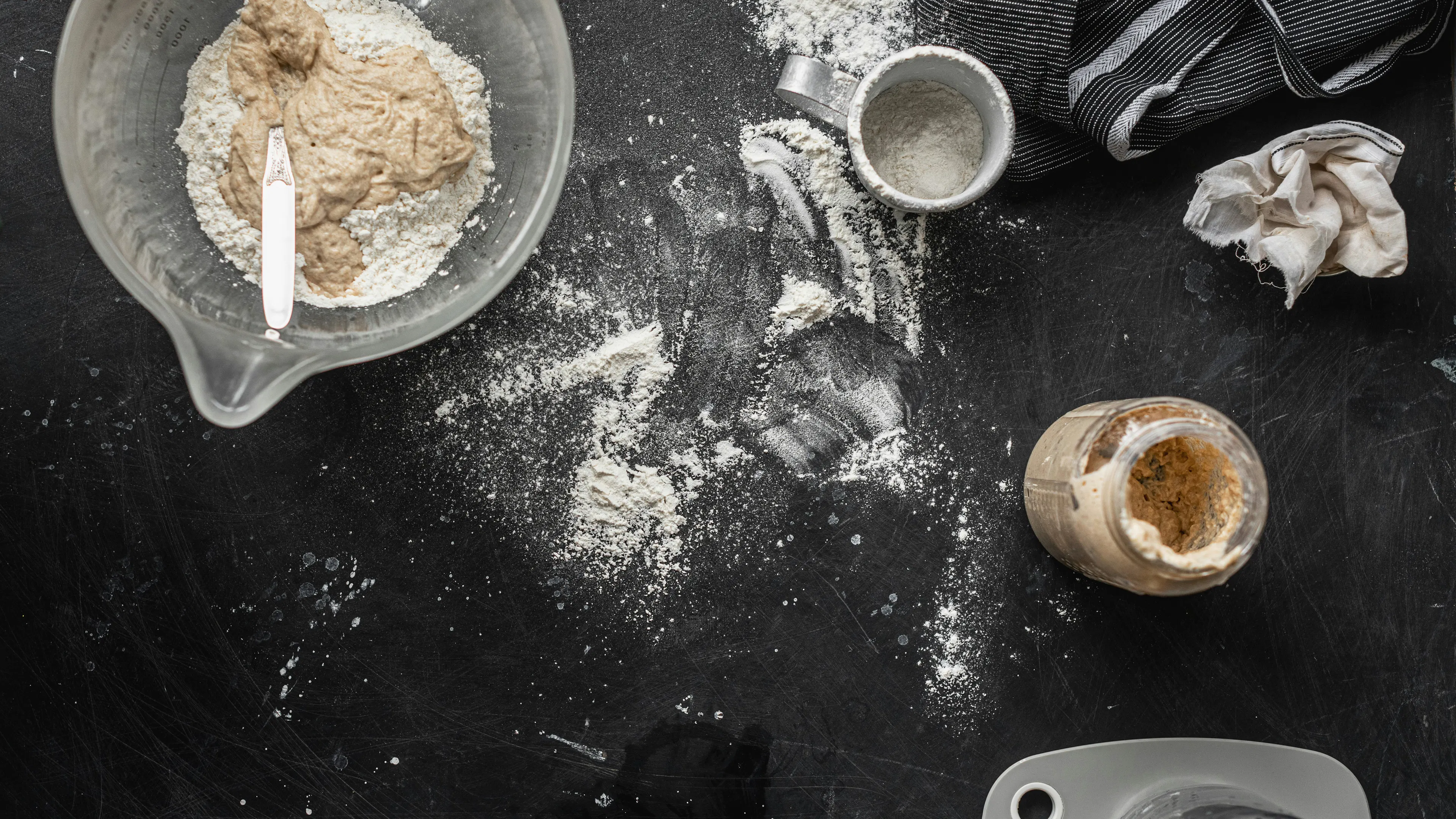 An overhead shot of baking ingredients and equipment on a dark, flour-dusted surface. A clear mixing bowl with dough and flour, a measuring cup, a jar of what appears to be sourdough starter, and a dishtowel are visible.