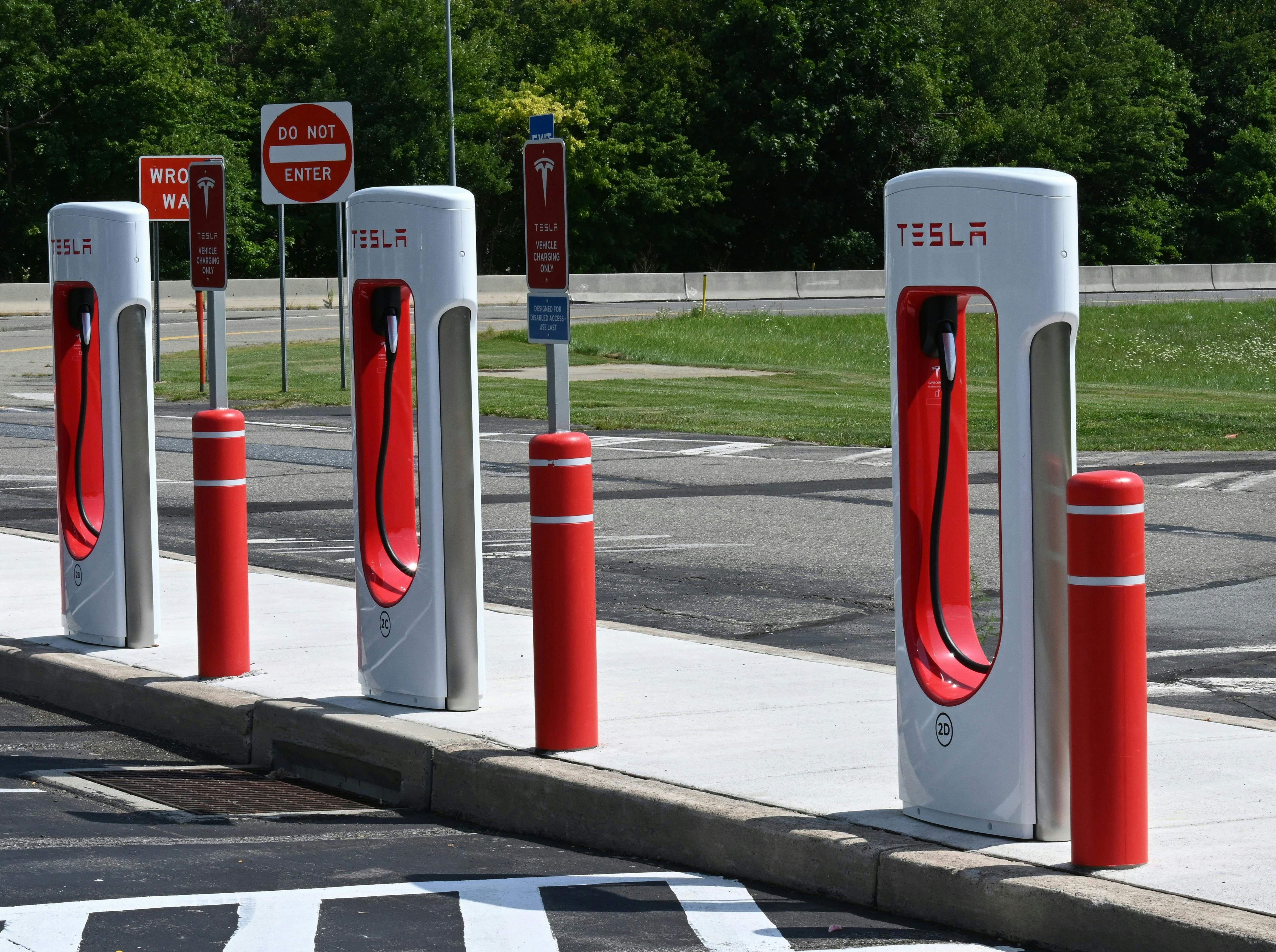 A row of four Tesla Supercharger stations, with white pillars and red accents, are lined up along a curb in a paved lot. "DO NOT ENTER" and "TESLA" signs are visible in the background, along with green trees under a clear sky.