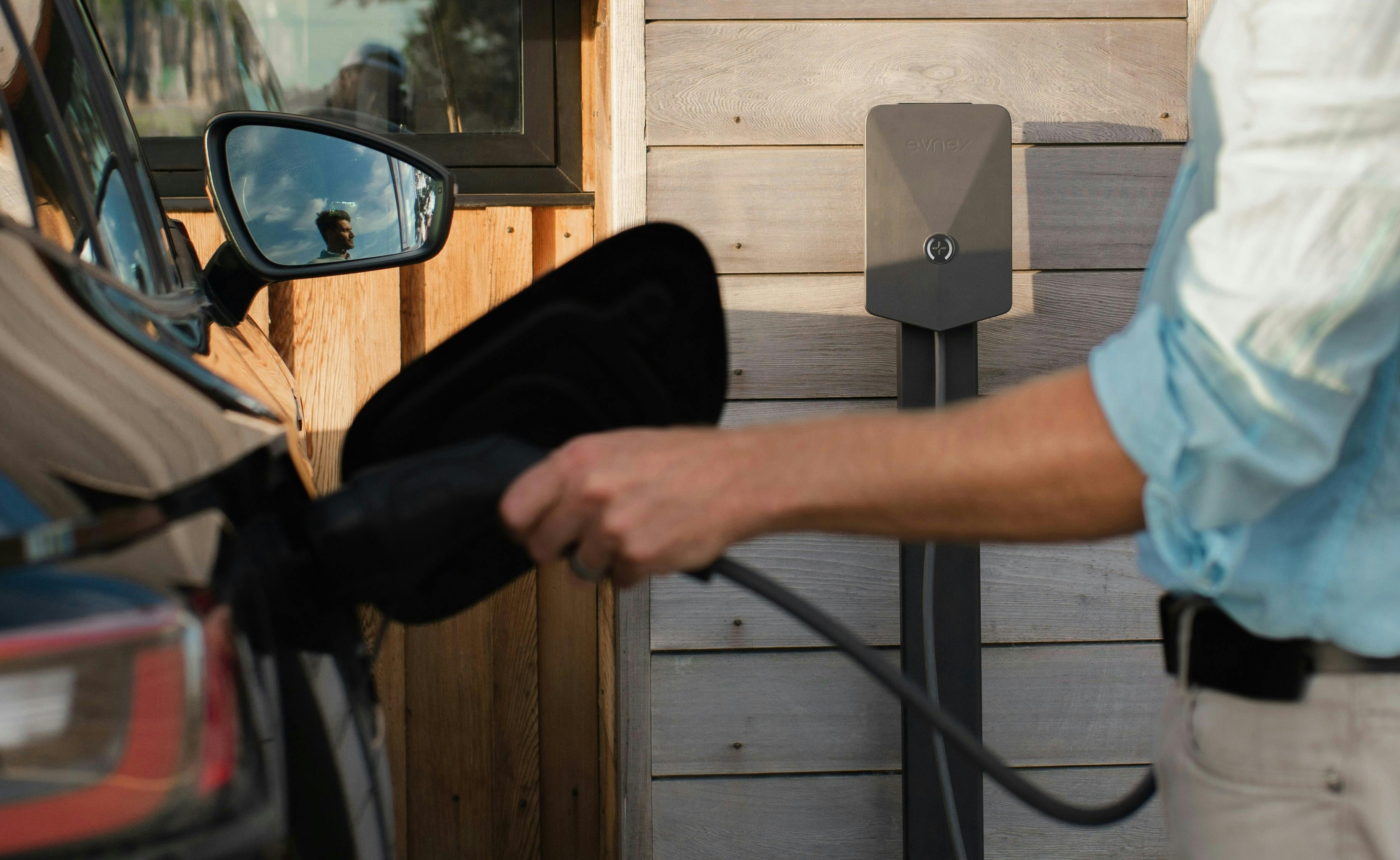 A person's arm is shown holding an EV charging cable and plugging it into the charging port of a dark-colored car. The car is parked next to a wooden-sided building with a grey wall-mounted charging unit.