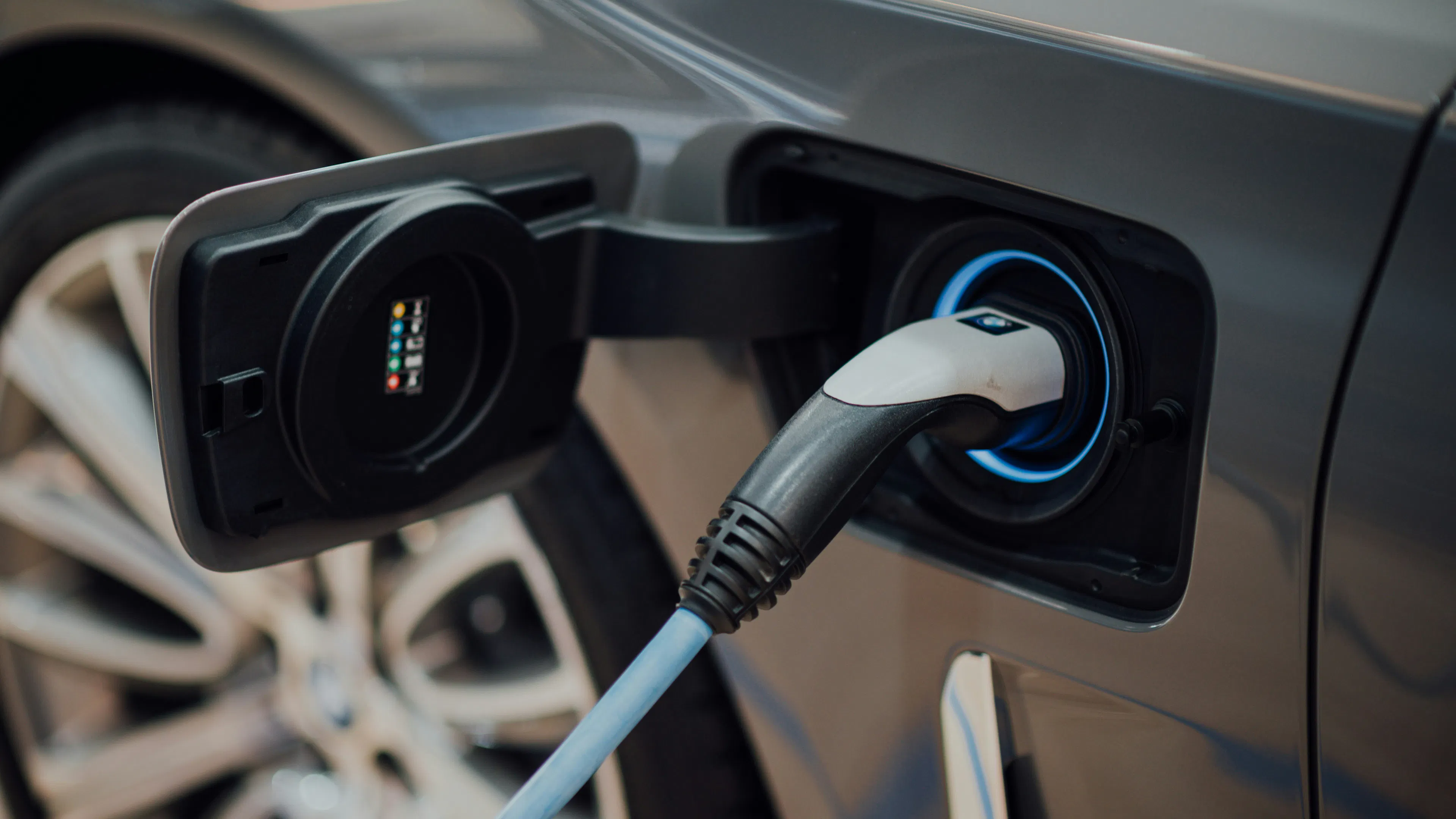 A close-up shot shows a charging cable plugged into the charge port of a grey electric vehicle. A blue light glows around the port, indicating it is actively charging. The car's wheel is partially visible in the background.