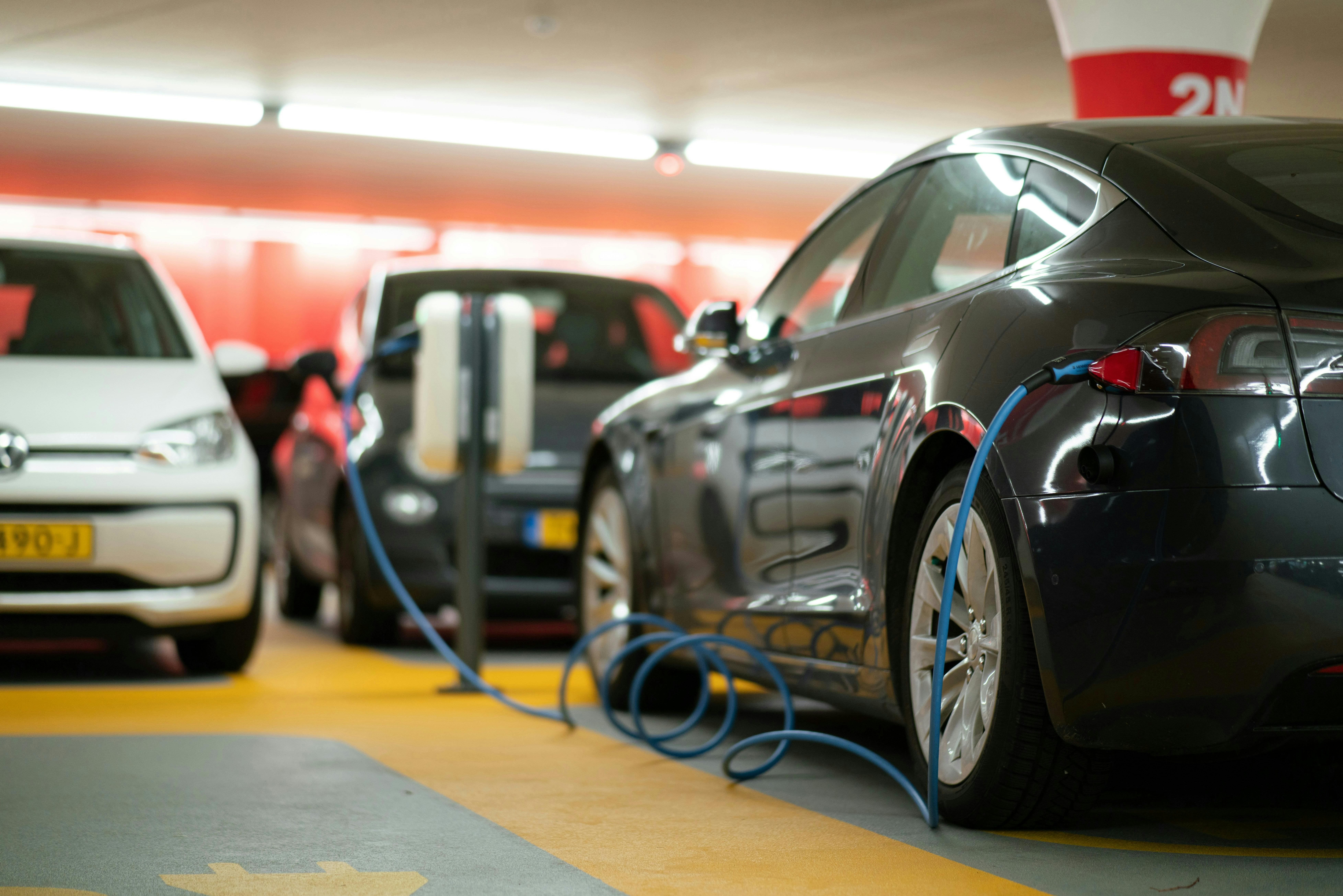 A dark grey Tesla Model S is charging in a parking garage, with a blue charging cable connected to its rear. A white Volkswagen is parked behind it, and red walls are visible in the background.