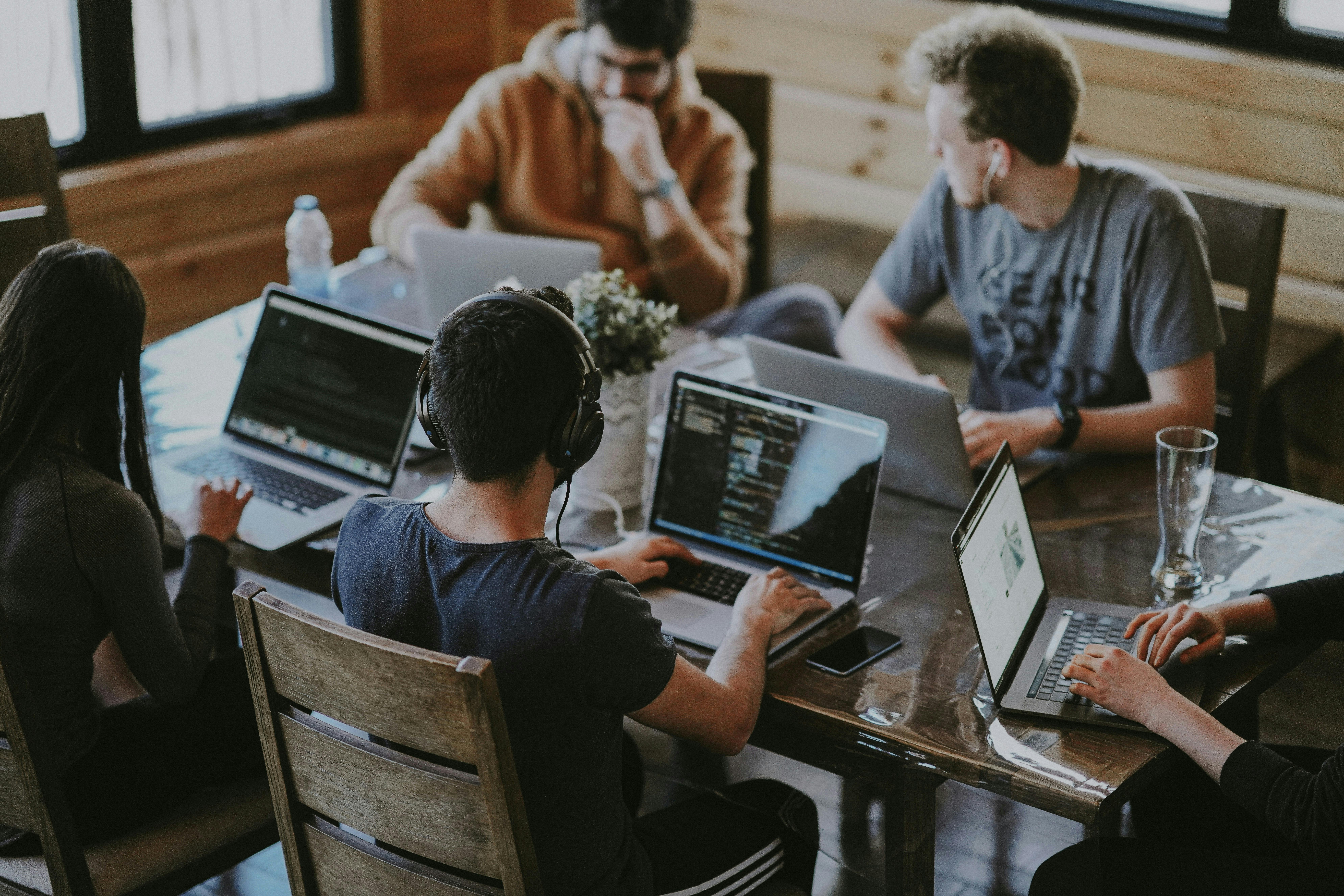 An overhead view of five people, likely students or collaborators, working on laptops around a large wooden table. Some are wearing headphones.