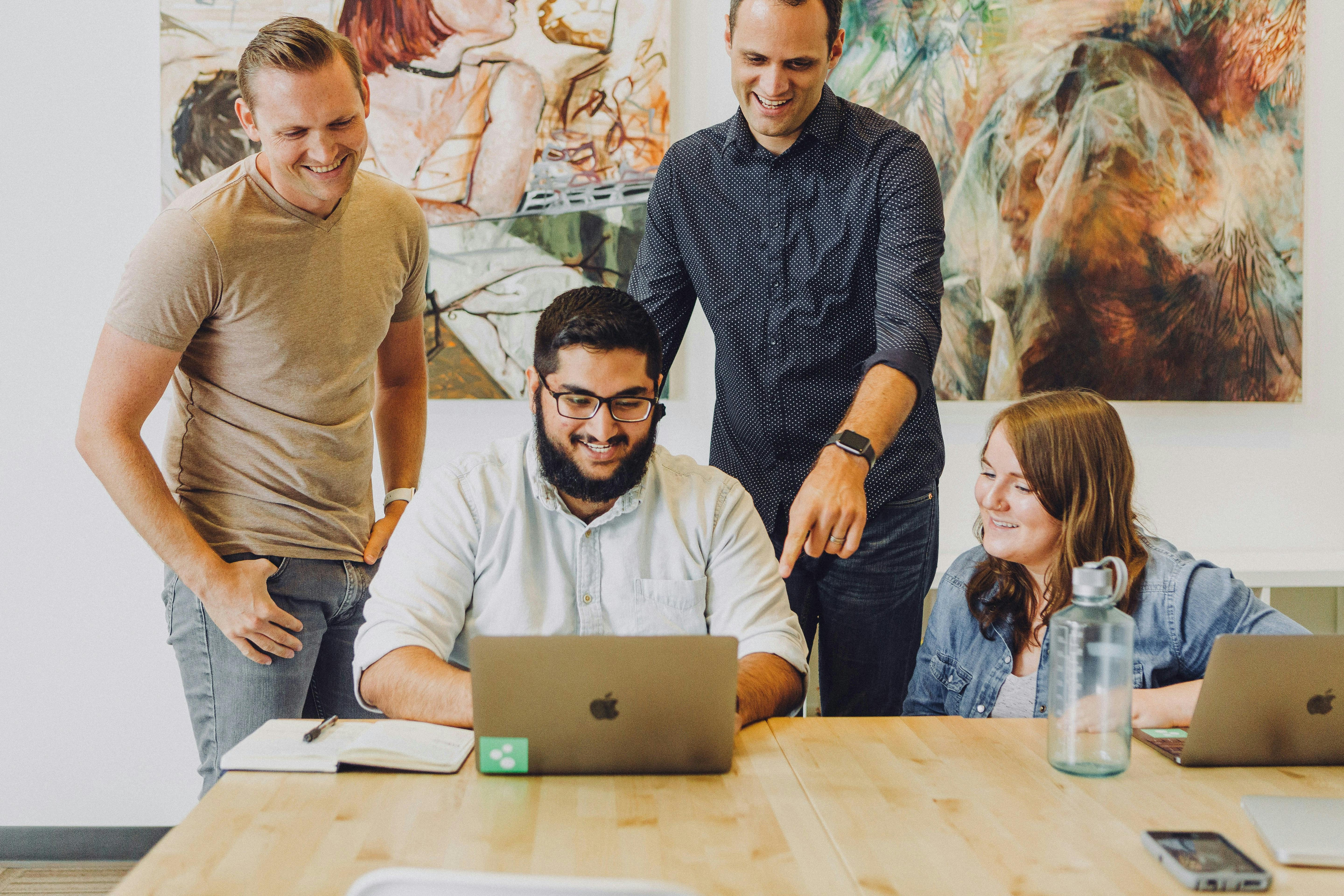 Four young adults are gathered around a wooden table with laptops. One man with glasses and a beard is seated at a laptop, with two men standing beside him and a woman seated to his right, all looking at the screen and smiling. Abstract art hangs on the wall in the background.