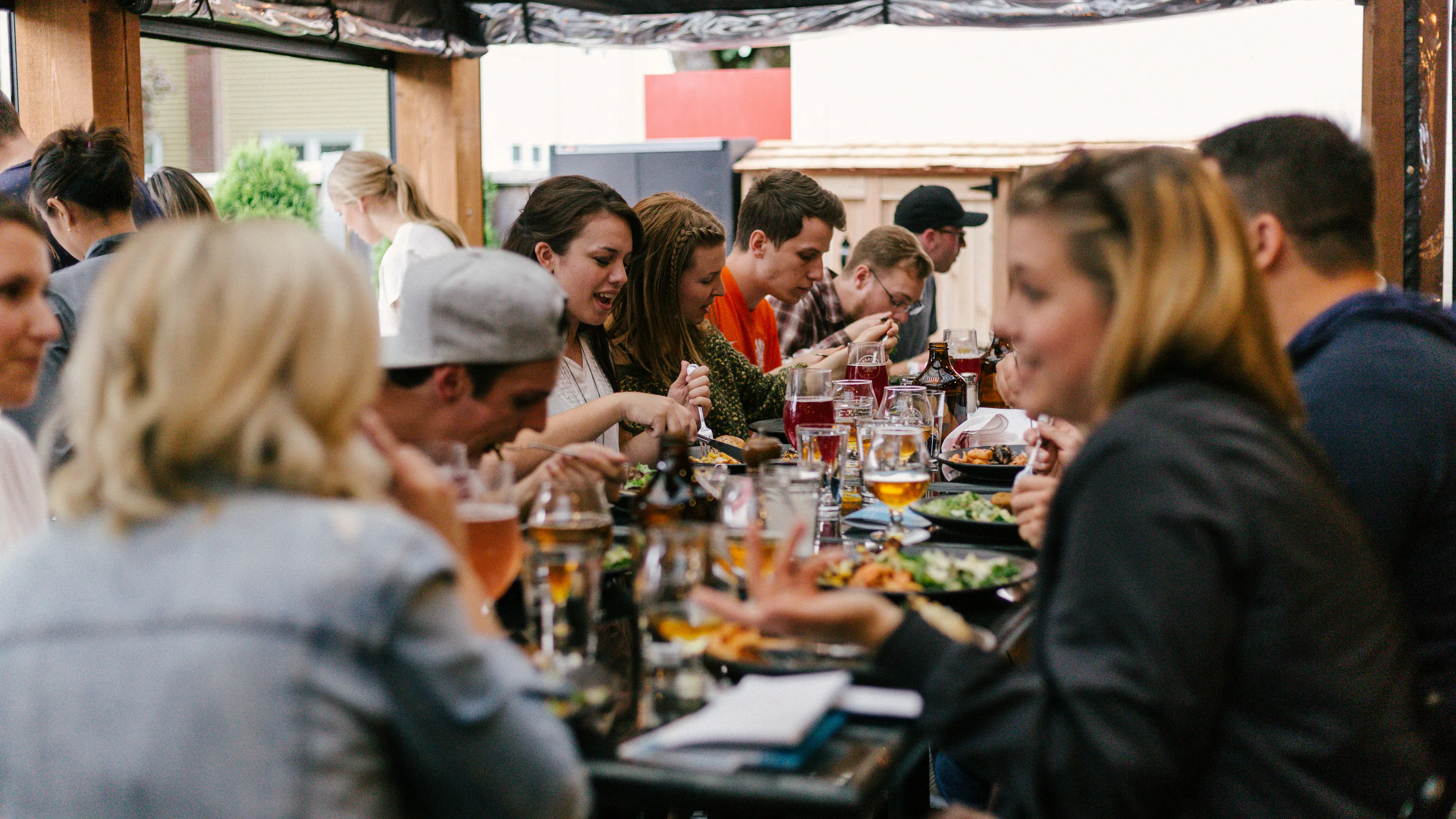 A diverse group of people are seated at a long outdoor table, enjoying a meal and drinks. Plates of food and glasses of beer are visible, and people are engaged in conversation. The scene suggests a lively social gathering.