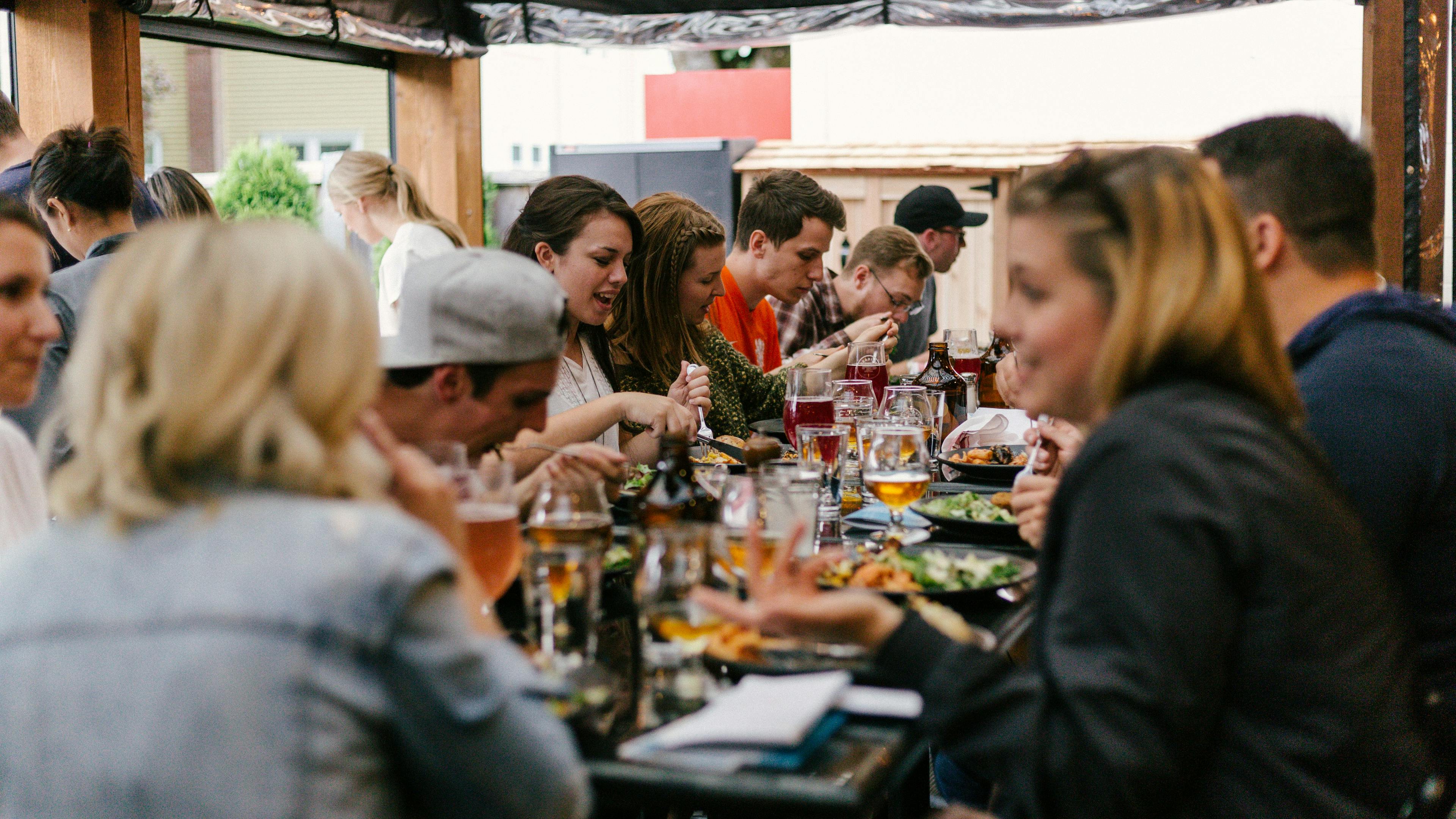 A diverse group of people are seated at a long outdoor table, enjoying a meal and drinks. Plates of food and glasses of beer are visible, and people are engaged in conversation. The scene suggests a lively social gathering.
