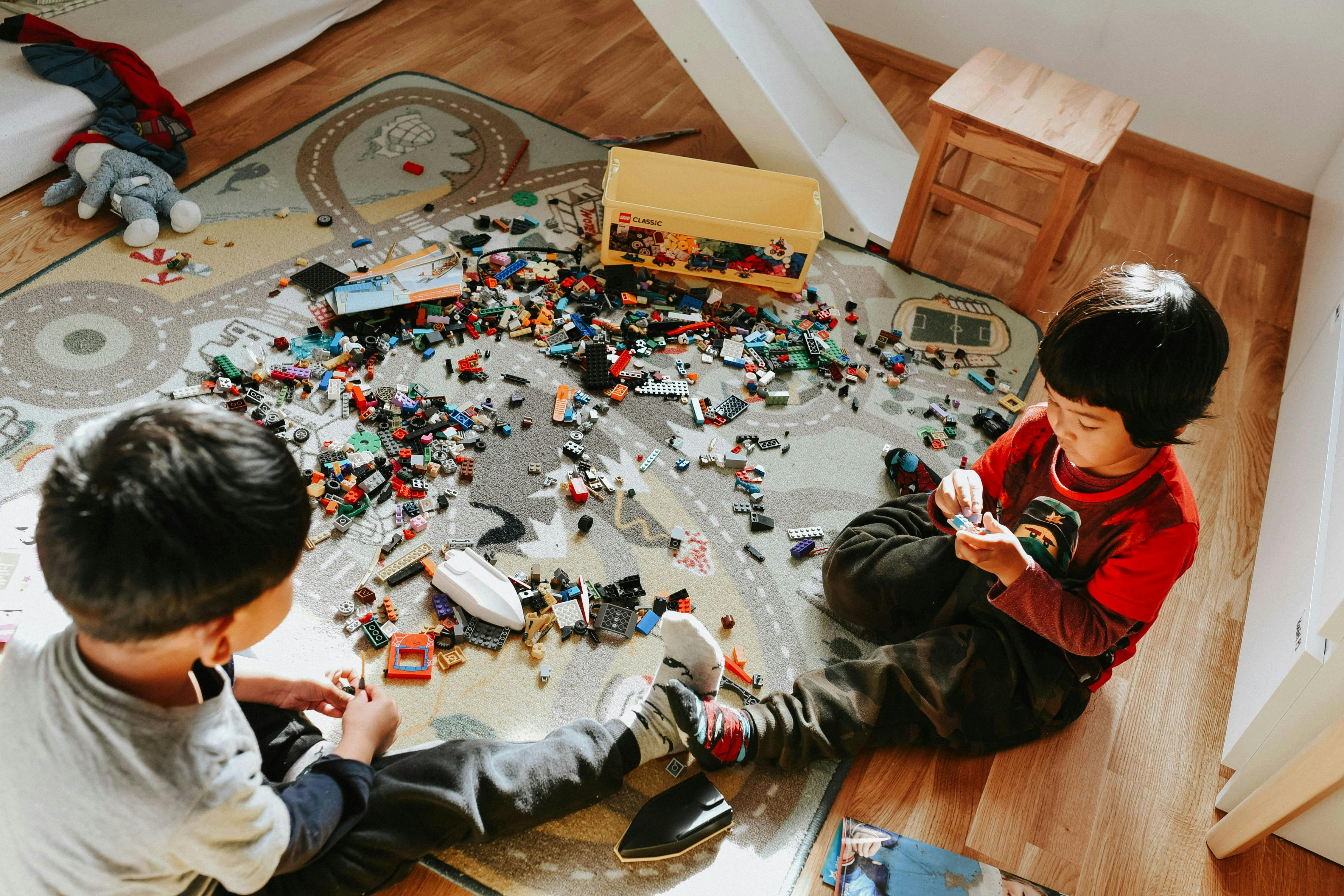 Two young children are sitting on a rug with a road map design, surrounded by a large pile of colorful building blocks and toys. One child is on the left facing away, and the other on the right is looking down at a toy.