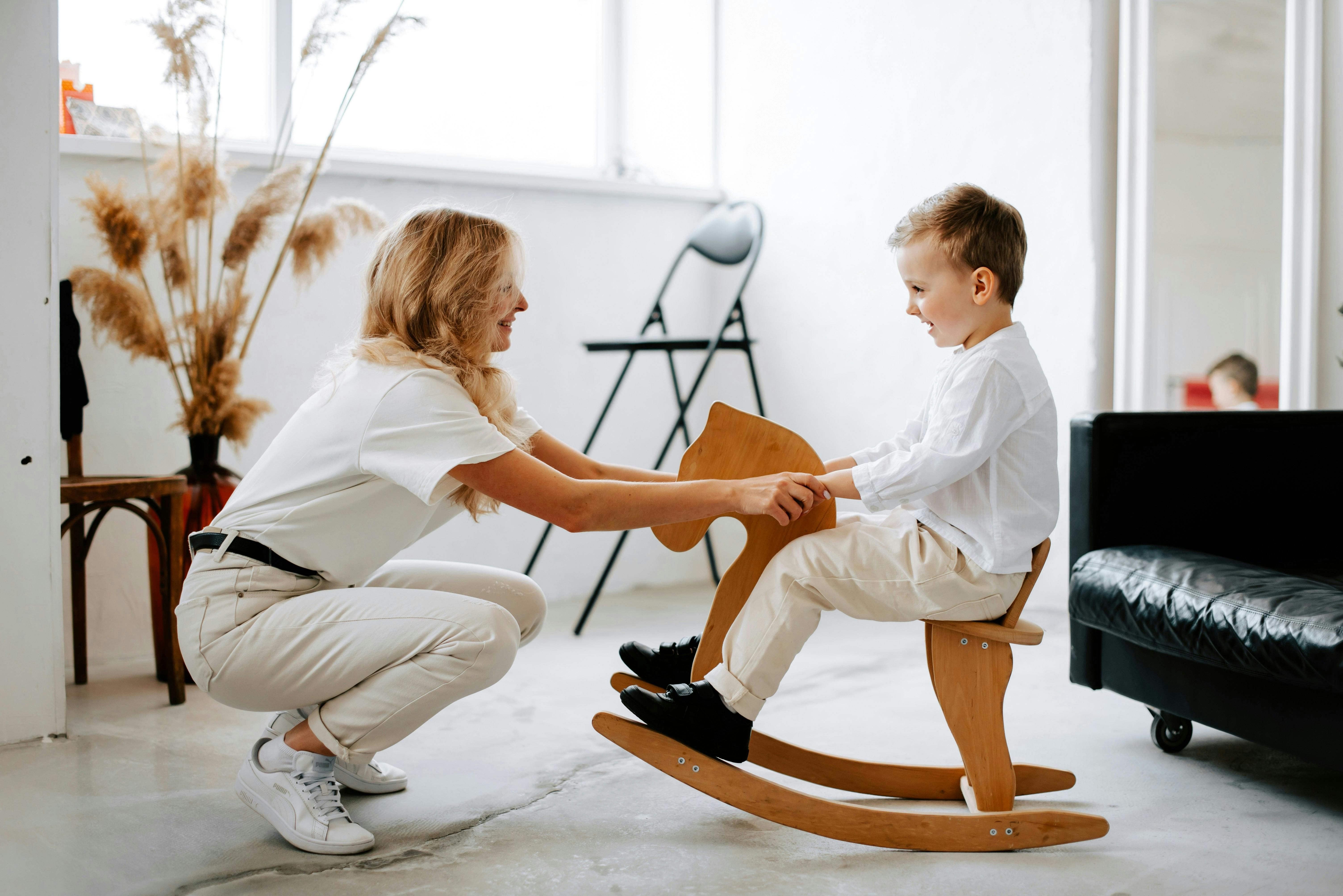 A woman with blonde hair, wearing a white shirt and light pants, crouches down, smiling at a young boy who is seated on a wooden rocking horse. They are in a brightly lit room with minimalist decor.