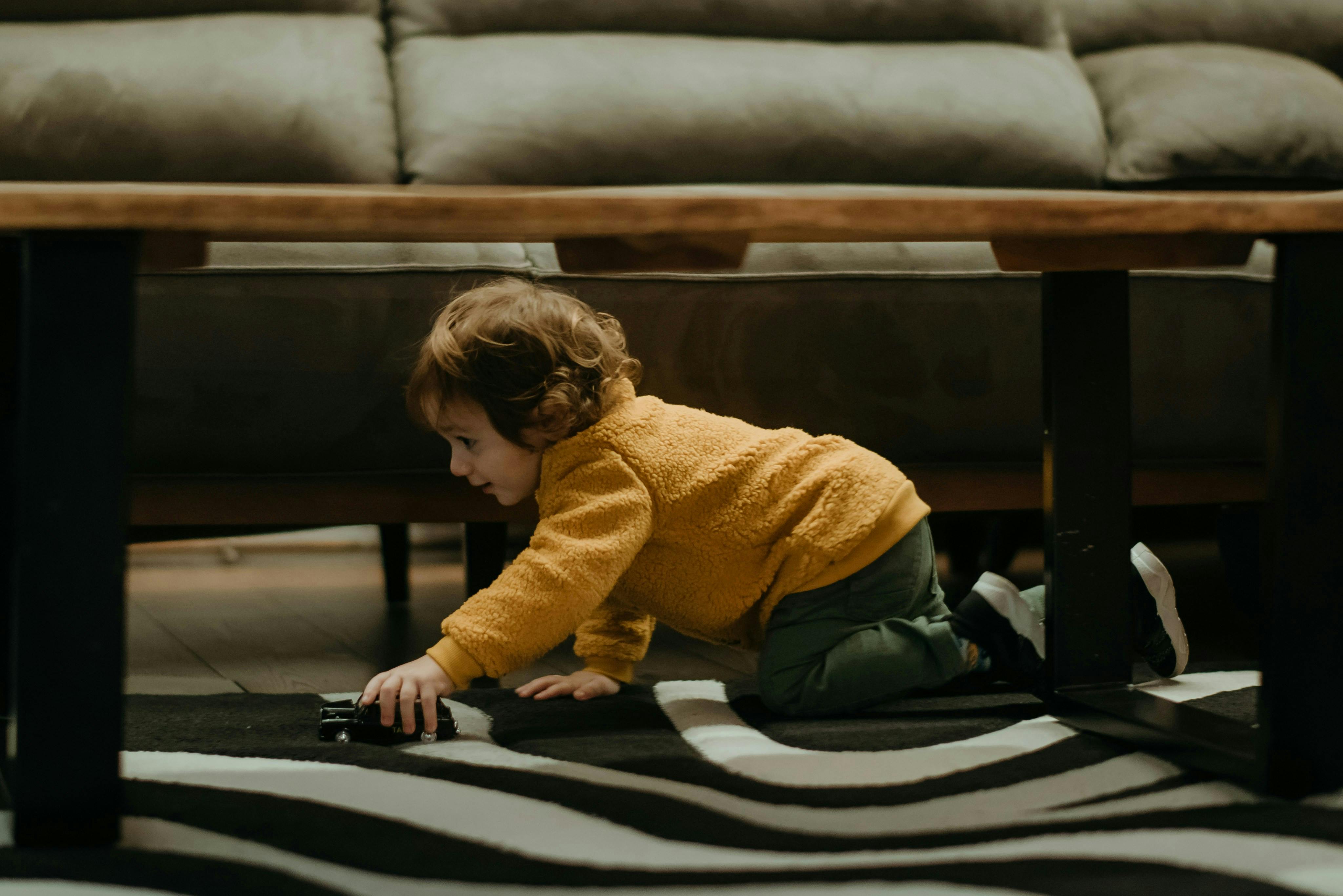 A young child with curly brown hair, wearing a yellow fuzzy top and green pants, is on their hands and knees under a coffee table, playing with a small toy car on a black and white striped rug. A sofa is visible in the background.
