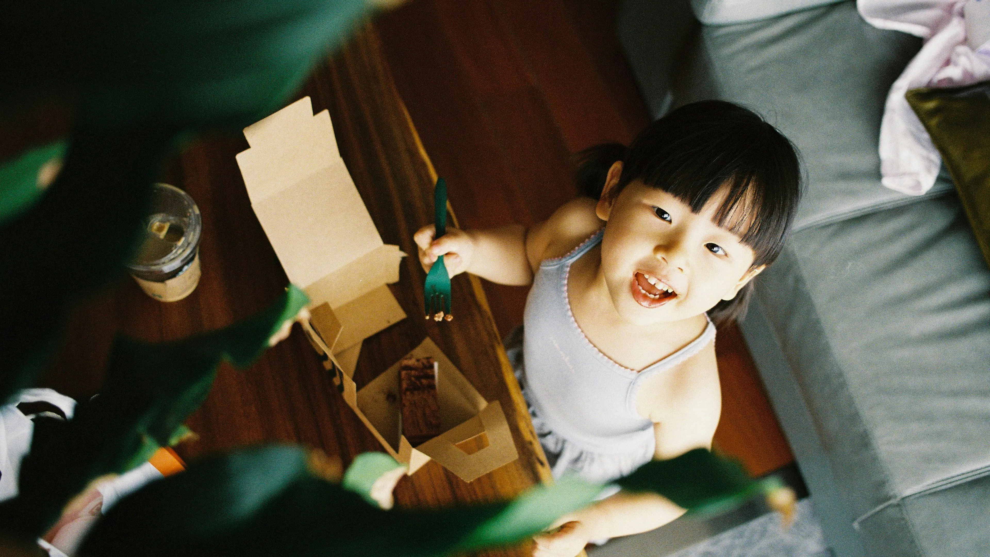 A young child with dark hair in pigtails, wearing a light-colored top, looks up at the camera with a wide smile, holding a green fork. They are next to an open cardboard takeout box on a wooden table, with a couch in the background.