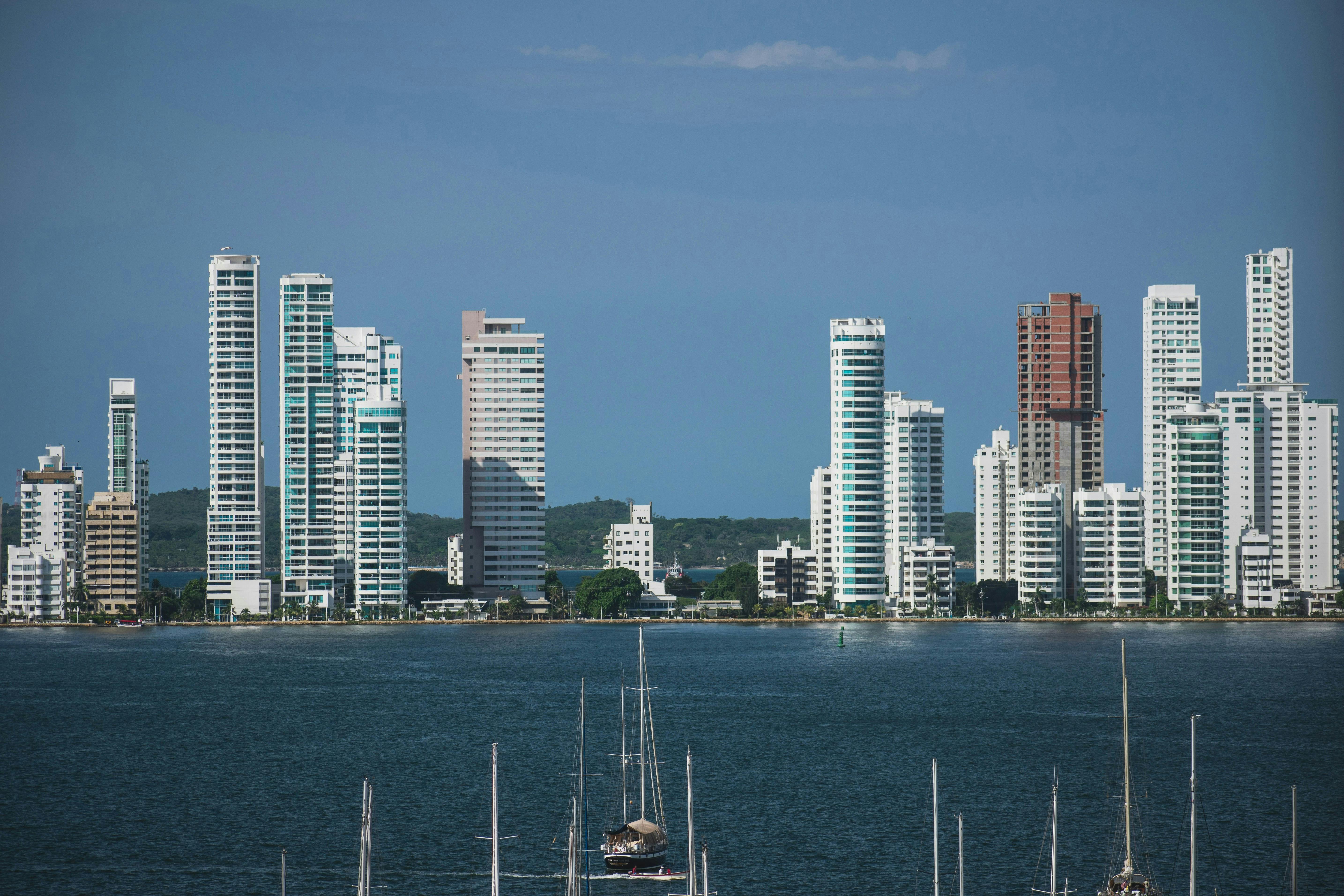 A cityscape with tall, modern buildings, some with blue glass, visible across a body of water. Several sailboats are docked or moored in the foreground. The sky is clear blue.
