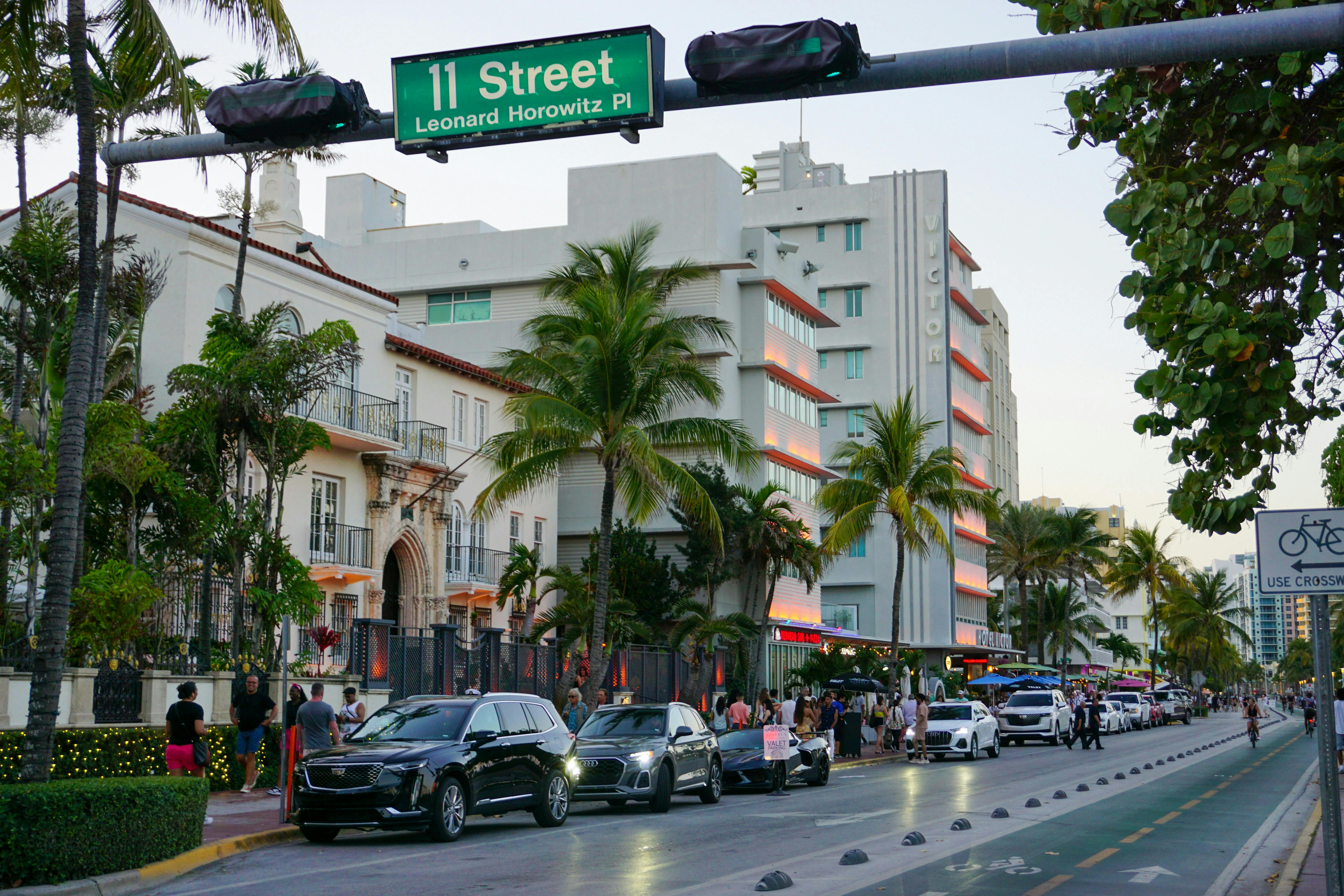 A vibrant street scene in Miami Beach, with Art Deco hotels and palm trees lining the street. Cars are parked along the curb, and people are walking on the sidewalk. An overhead sign reads "11 Street Leonard Horowitz Pl."
