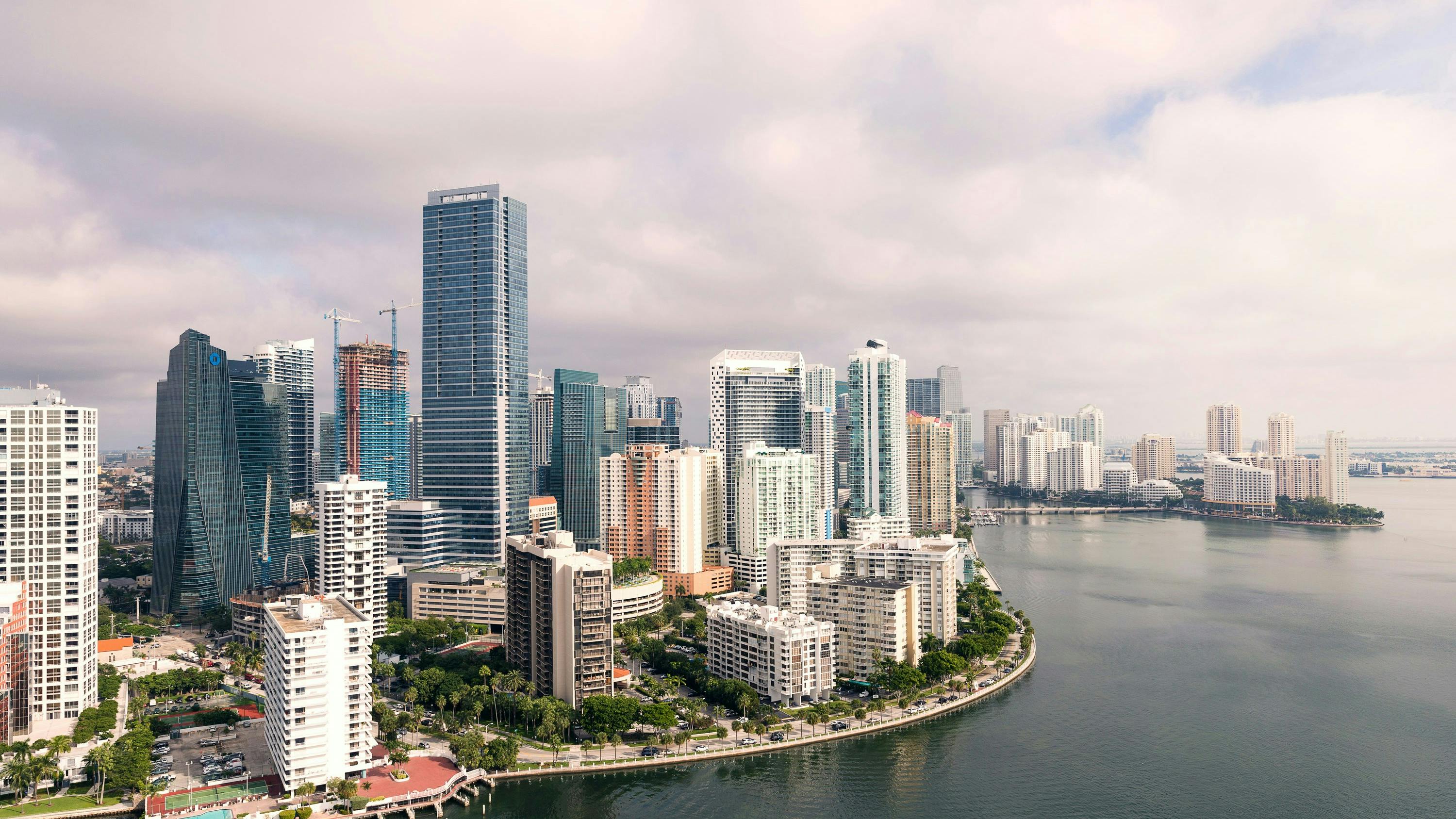 An aerial view of the Miami skyline on a cloudy day, showing numerous high-rise buildings along a curving waterfront.