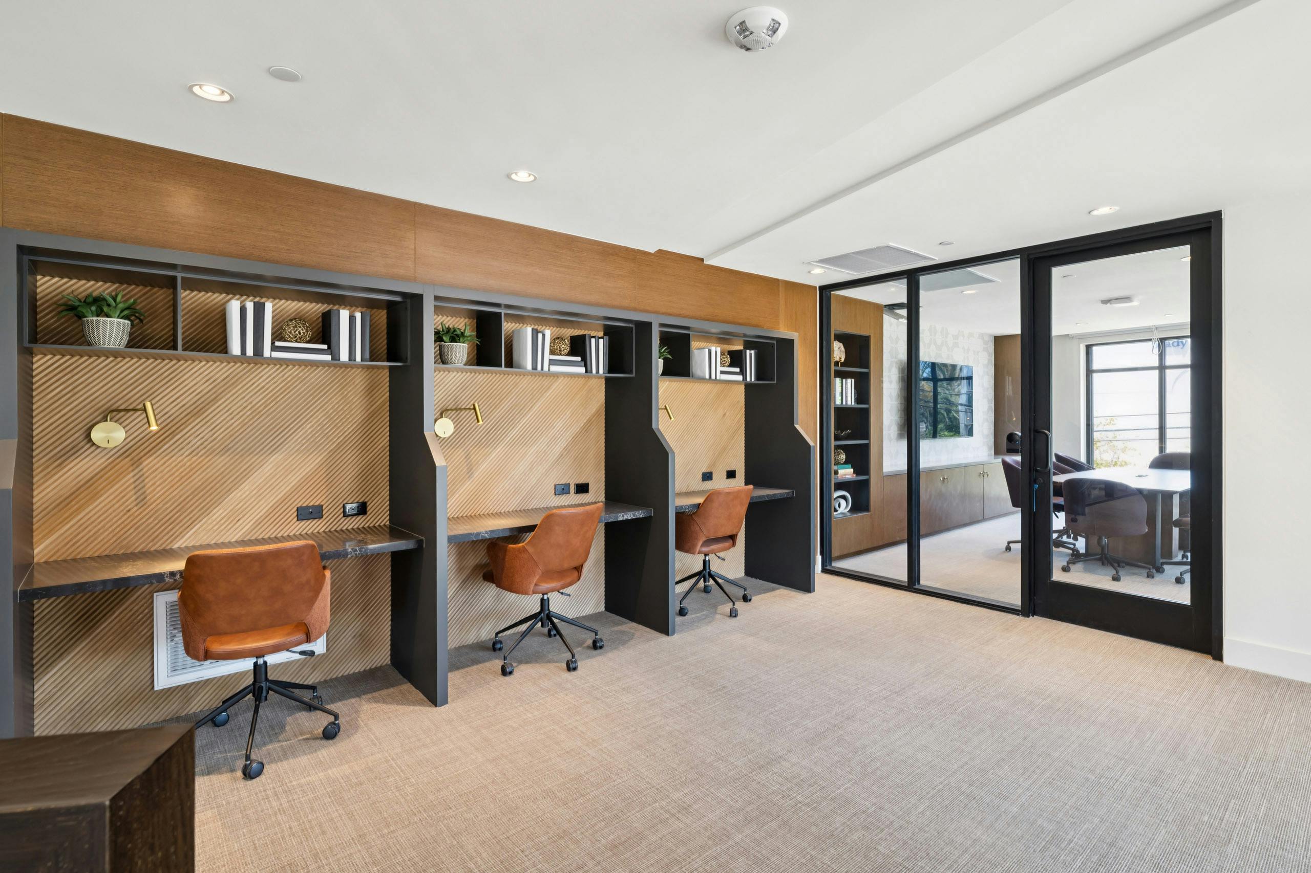Coworking area at AMLI Aero with built-in desks, warm wood accents, leather chairs, shelving, and glass wall to private conference room.