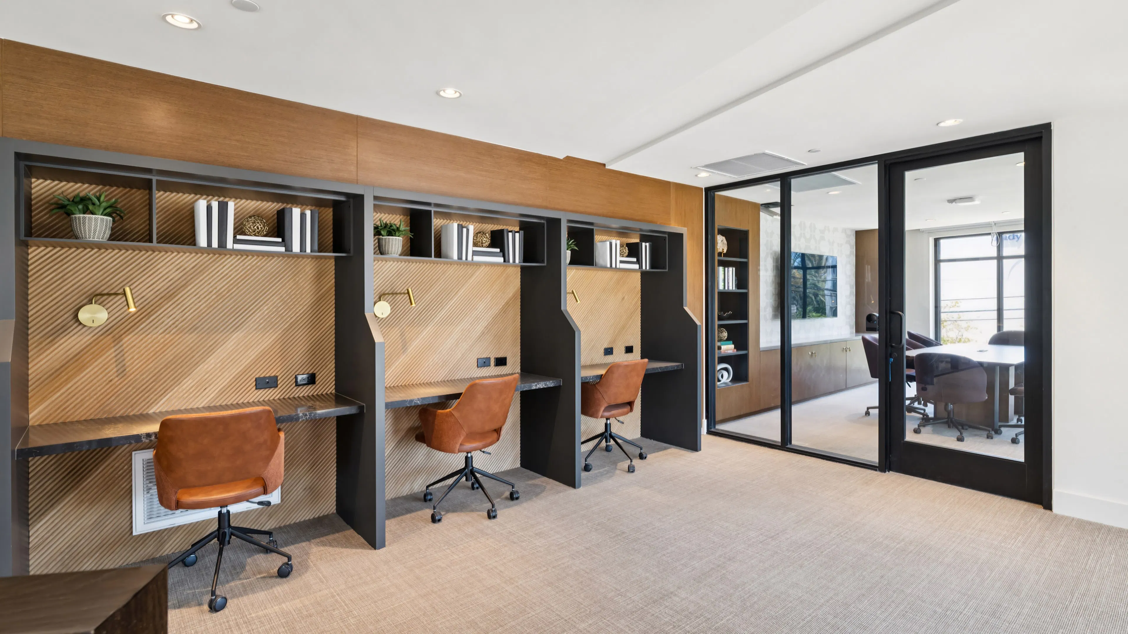 Coworking area at AMLI Aero with built-in desks, warm wood accents, leather chairs, shelving, and glass wall to private conference room.